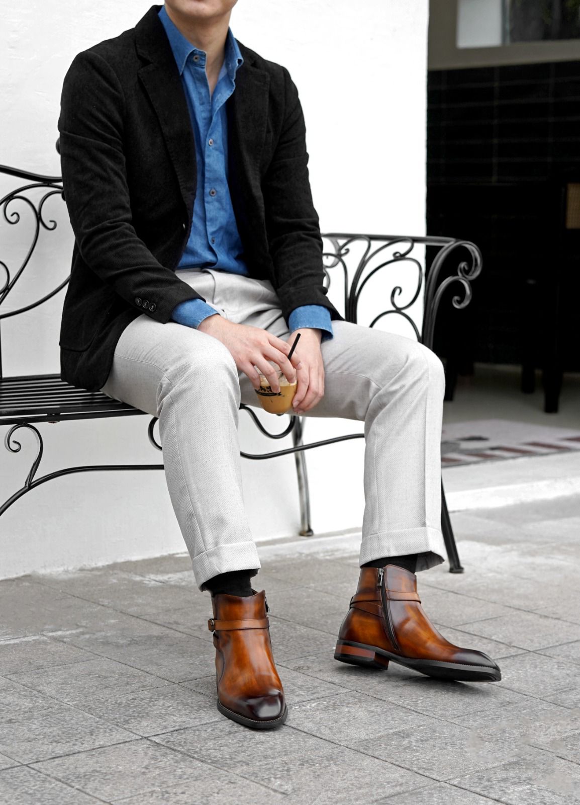 A man sits on a black wrought-iron bench outdoors, wearing a black blazer, blue button-up shirt, light gray trousers, and brown leather ankle boots.