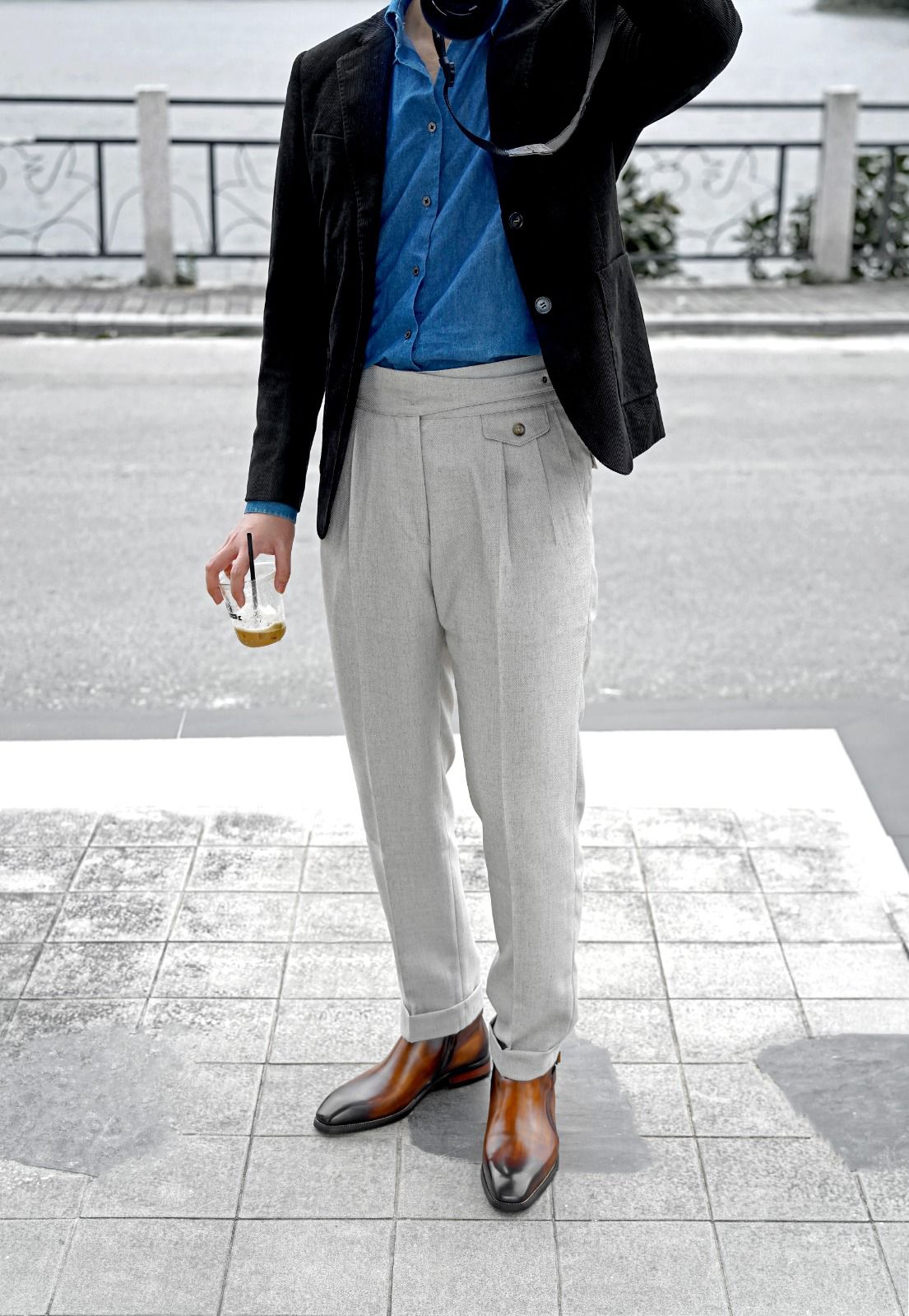 A man wearing a black velvet blazer, a bright blue button-down shirt, light gray pleated trousers, and brown leather cap-toe shoes, standing outdoors on a paved street.