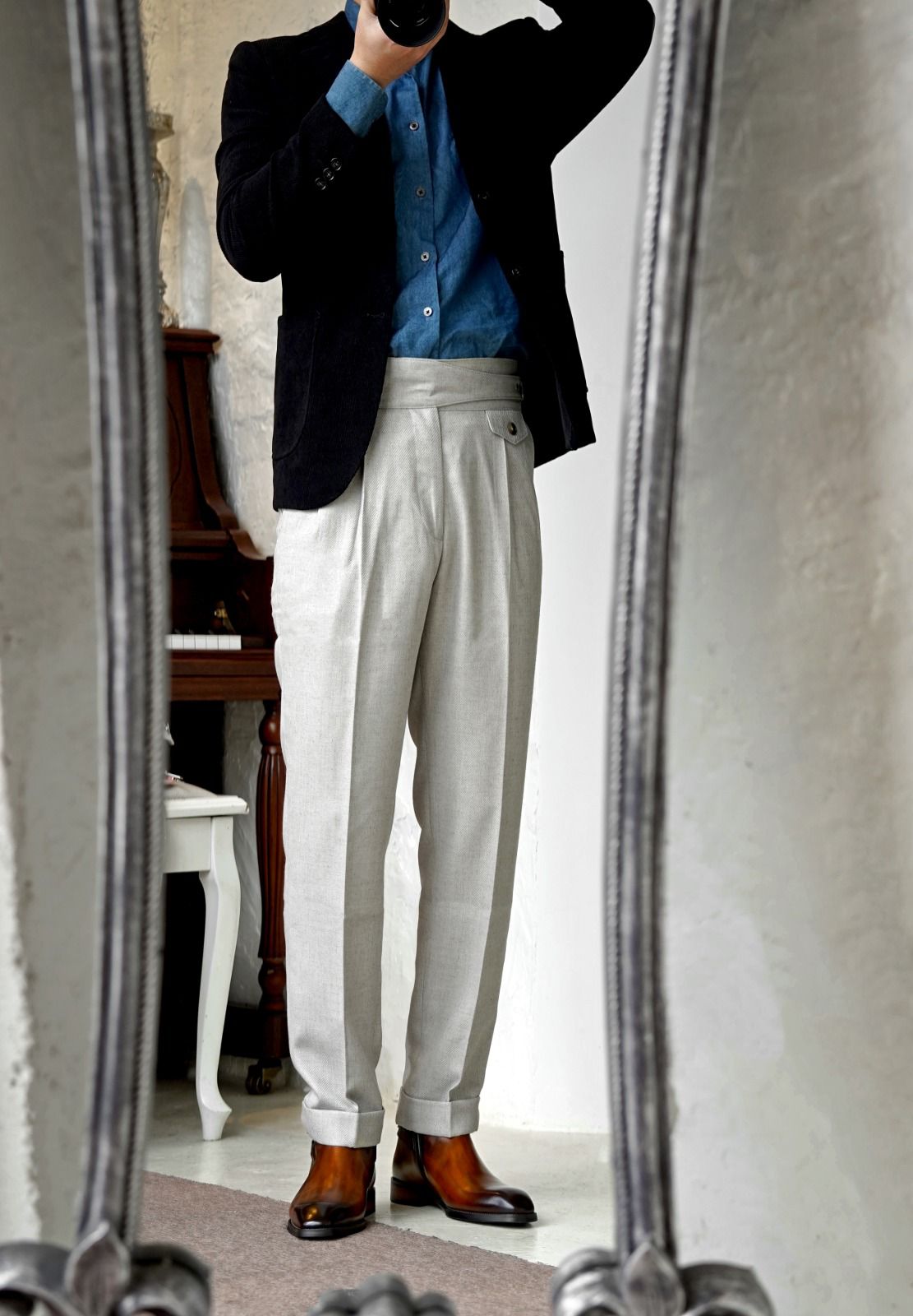 A man takes a mirror selfie wearing a dark navy blazer, a blue button-down shirt, light beige pleated trousers, and brown leather dress shoes in an indoor setting.
