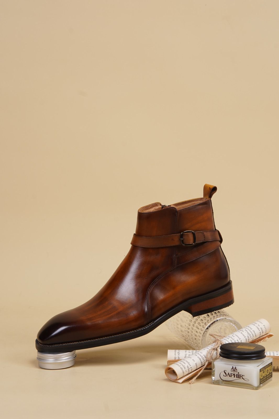 A brown leather ankle boot with a buckle strap, displayed on a metal stand with a small jar of Caviar cream and rolled papers on a beige background.
