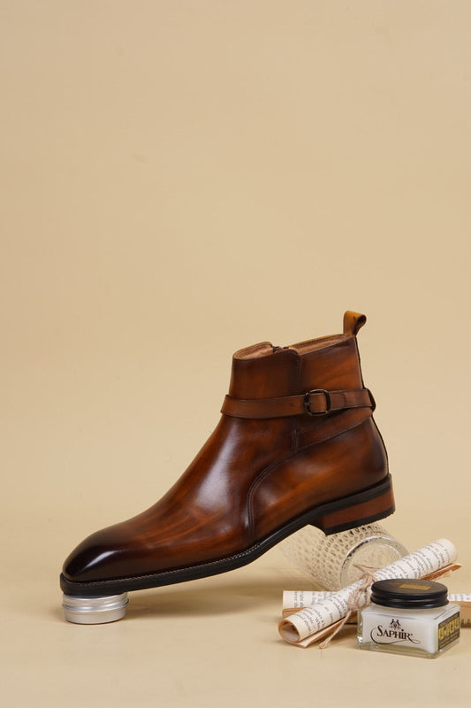 A brown leather ankle boot with a buckle strap, displayed on a metal stand with a small jar of Caviar cream and rolled papers on a beige background.