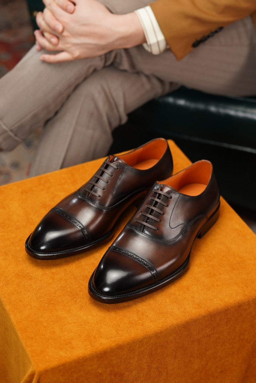 A pair of dark brown cap-toe oxford shoes with polished leather and lace-up fronts, displayed on an orange velvet-covered box with a person's legs and hands visible in the background.