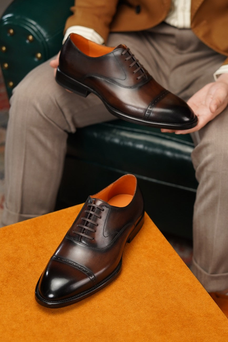 A pair of dark brown leather lace-up dress shoes with a cap toe, shown with one shoe held by a person sitting on a dark green leather chair and the other resting on an orange surface.