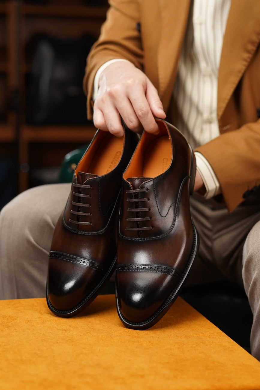 A pair of dark brown leather cap-toe oxford shoes held by a person wearing a tan blazer and striped shirt, sitting on a wooden surface with a warm orange-toned background.