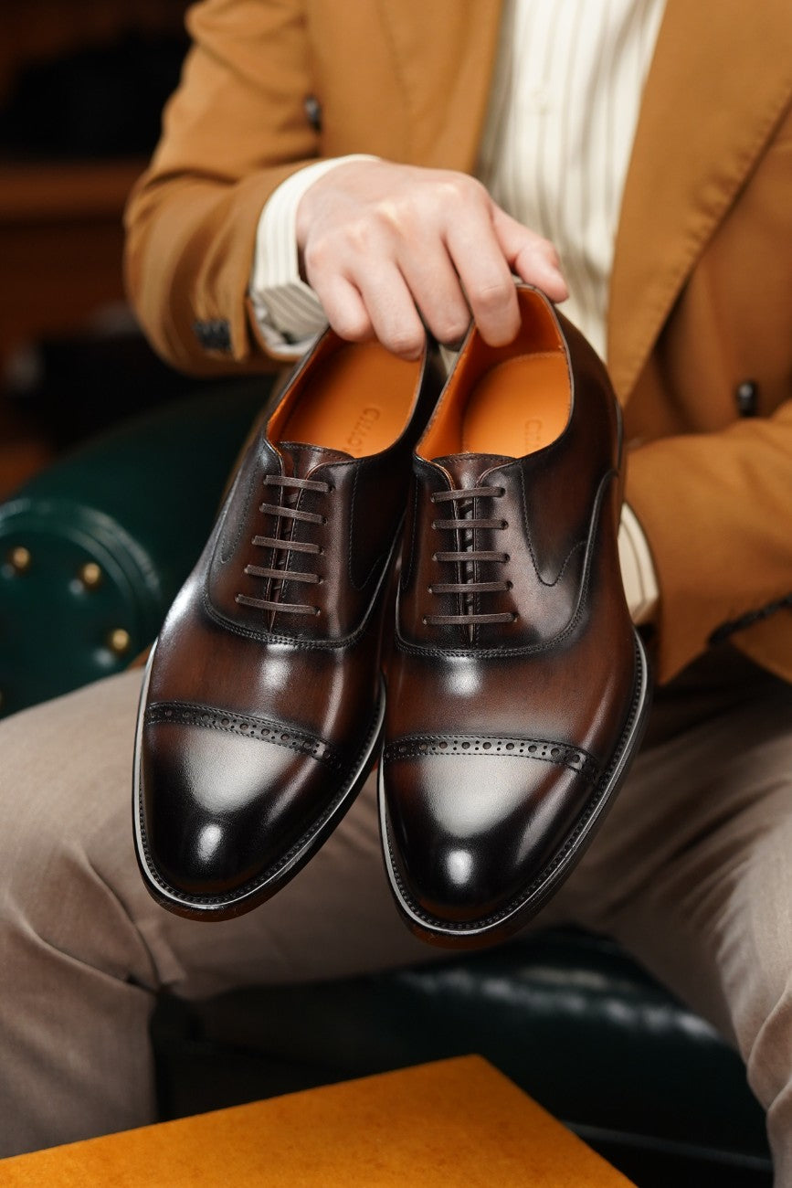 A person in a tan blazer holds a pair of dark brown leather cap-toe oxford shoes with brogue detailing and laces, photographed in a styled indoor setting.