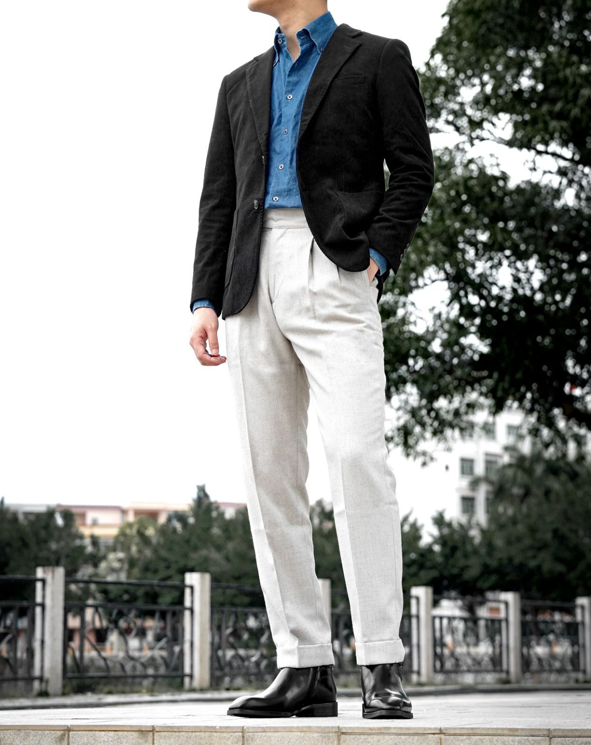 A man wearing a black blazer, blue button-down shirt, off-white trousers, and black leather shoes, standing outdoors on a stone ledge with trees and a railing in the background.