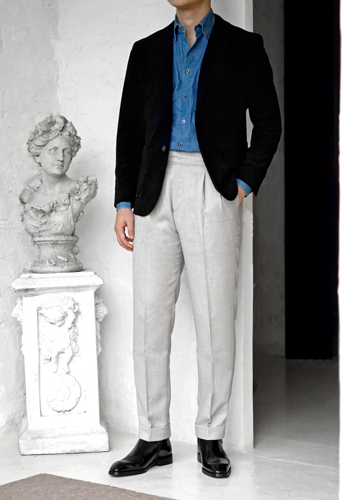 A man wearing a black velvet blazer, a blue button-down shirt, light gray pleated trousers, and black leather dress shoes, standing in a bright interior next to a white classical bust statue.