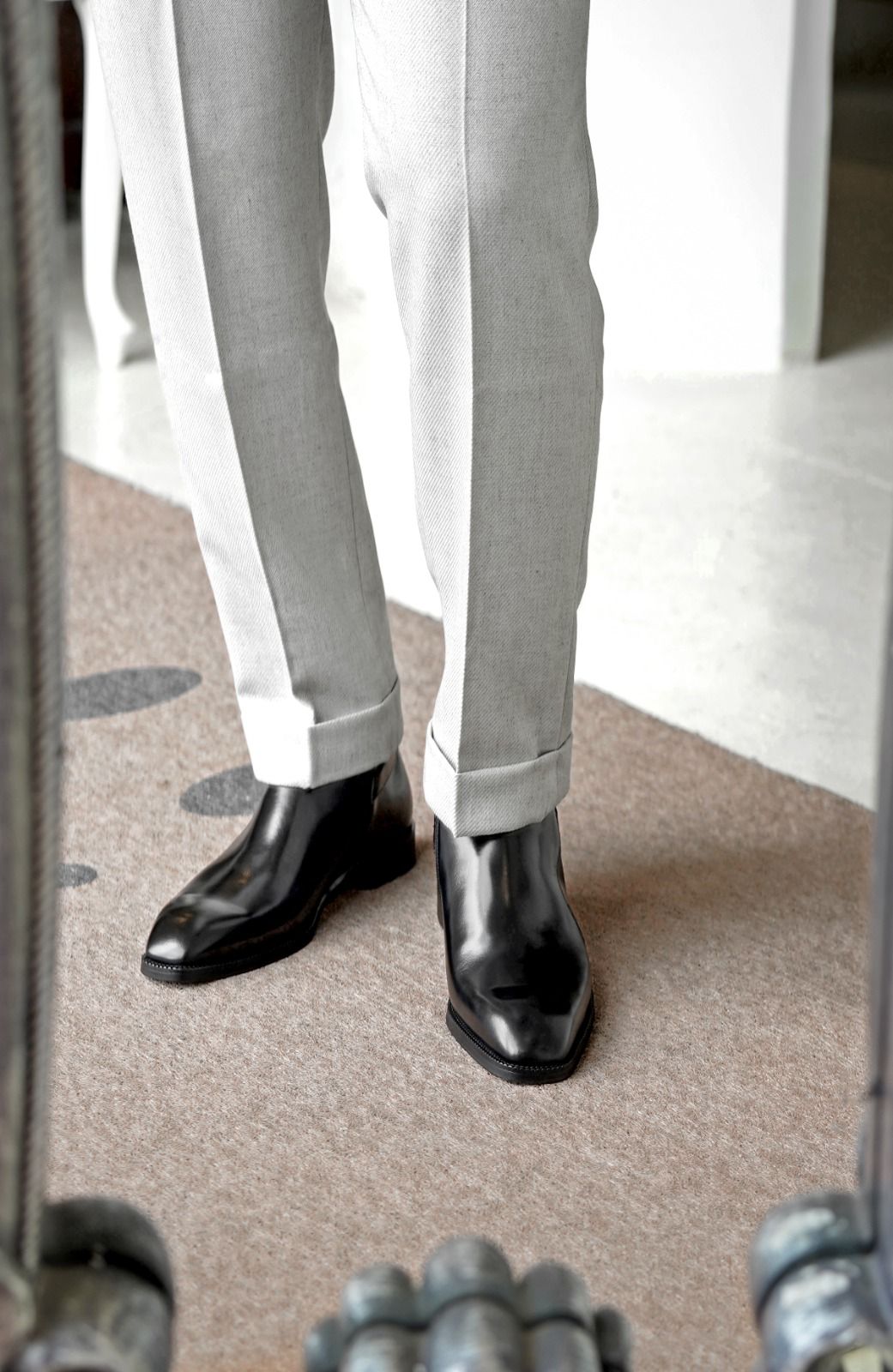 A close-up of a person's legs wearing light gray dress pants and shiny black leather boots, standing on a textured beige carpet.