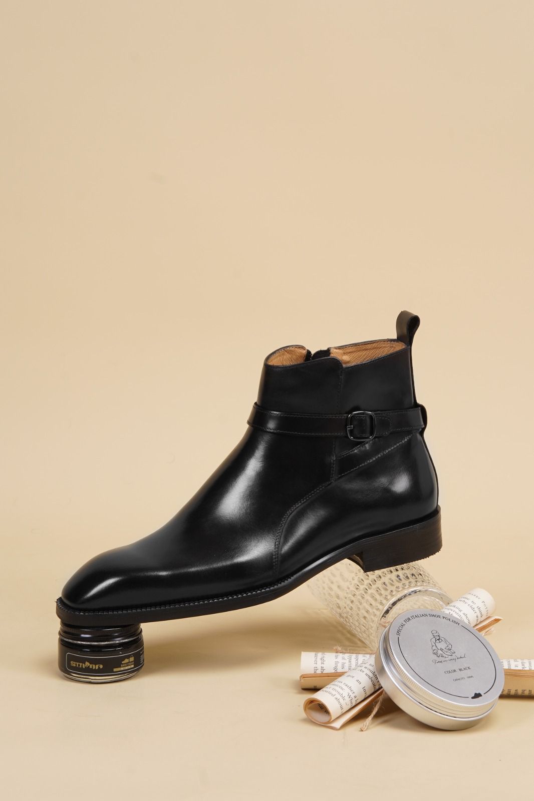 A black leather ankle boot with a buckle strap, displayed on a stack of books and a tin of shoe polish on a beige background.