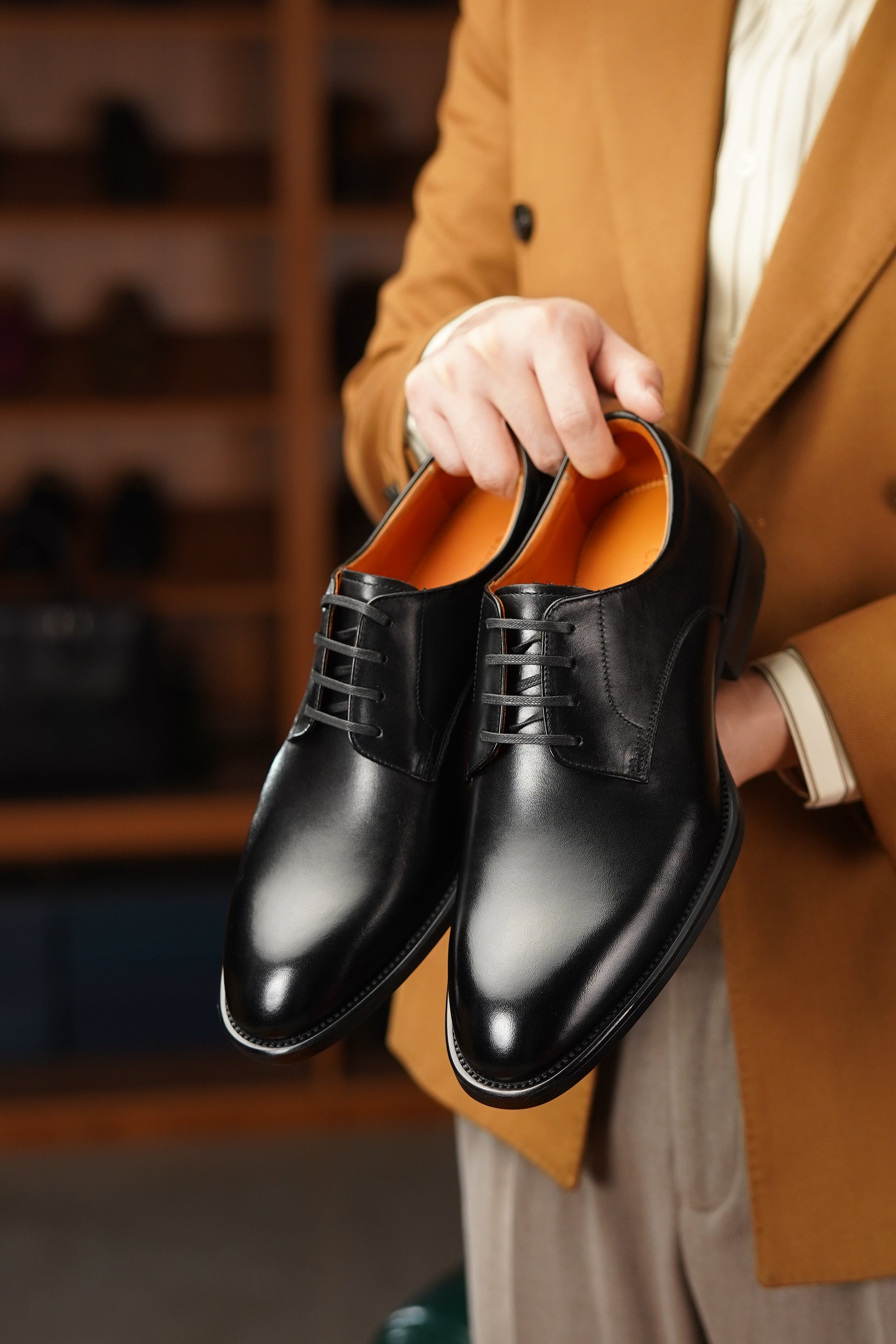 Person holding a pair of black leather shoes with an orange interior, wearing a brown coat.