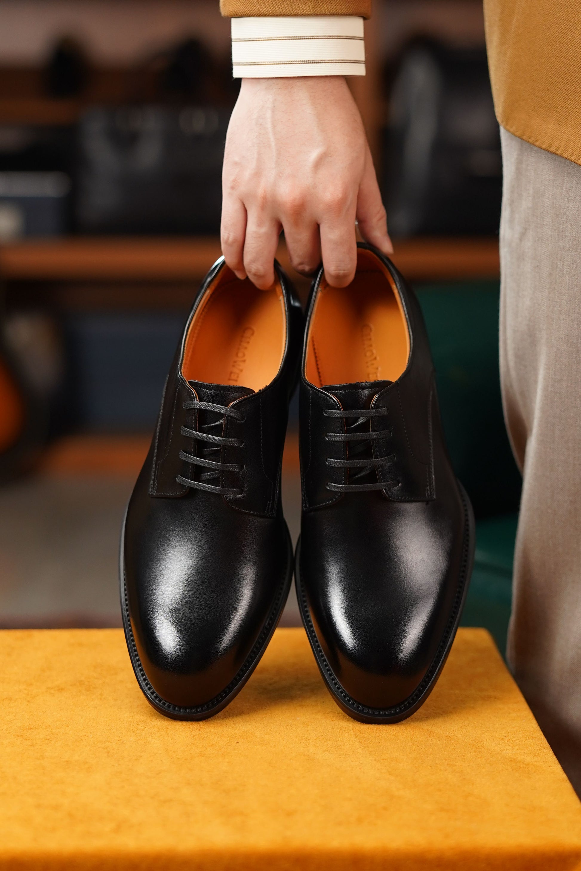 A pair of black leather lace-up dress shoes held by a person's hand over a mustard-yellow surface, with a blurred indoor background and part of the person's clothing visible.
