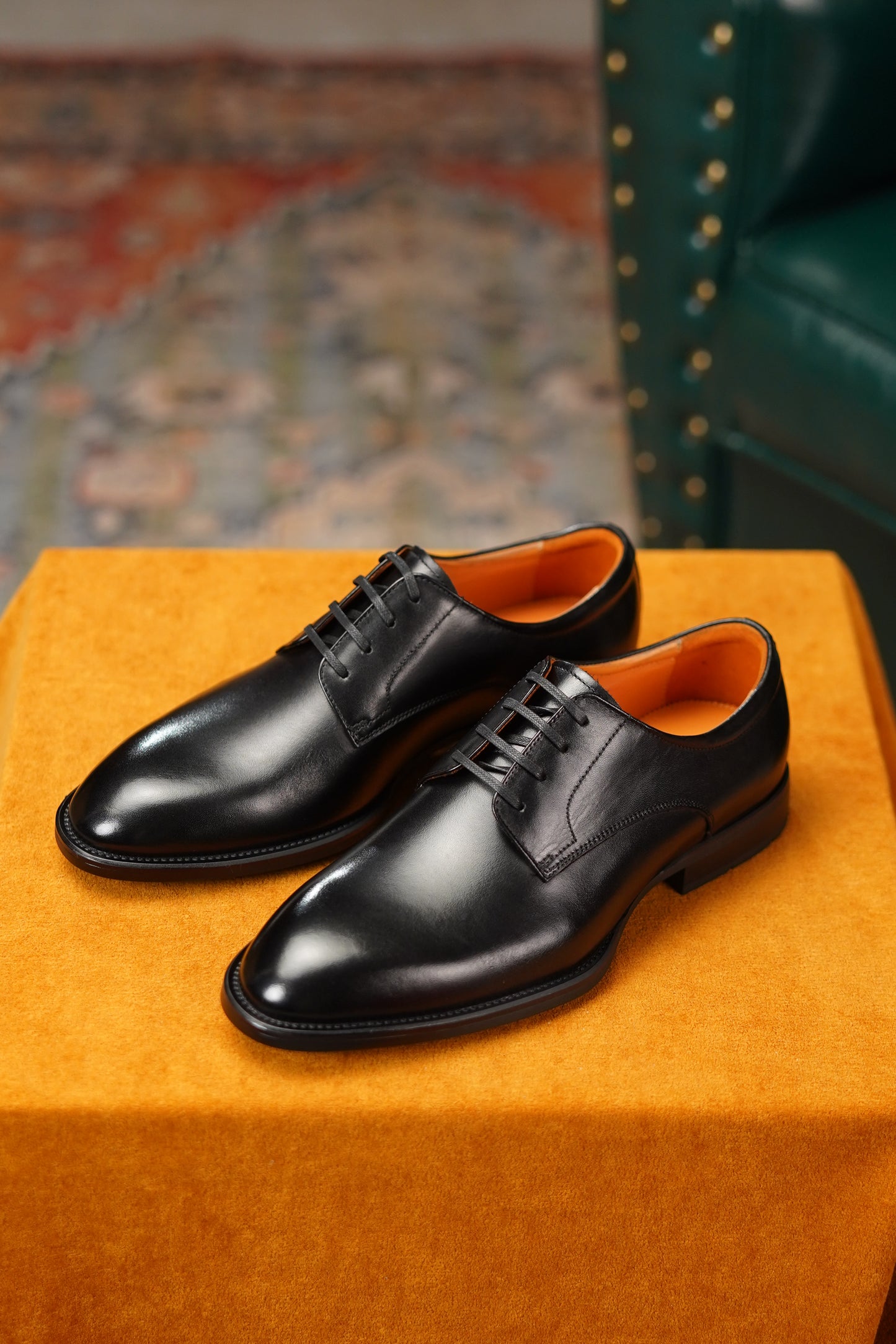 Pair of black dress shoes on an orange cushion with a patterned rug and green chair in the background.