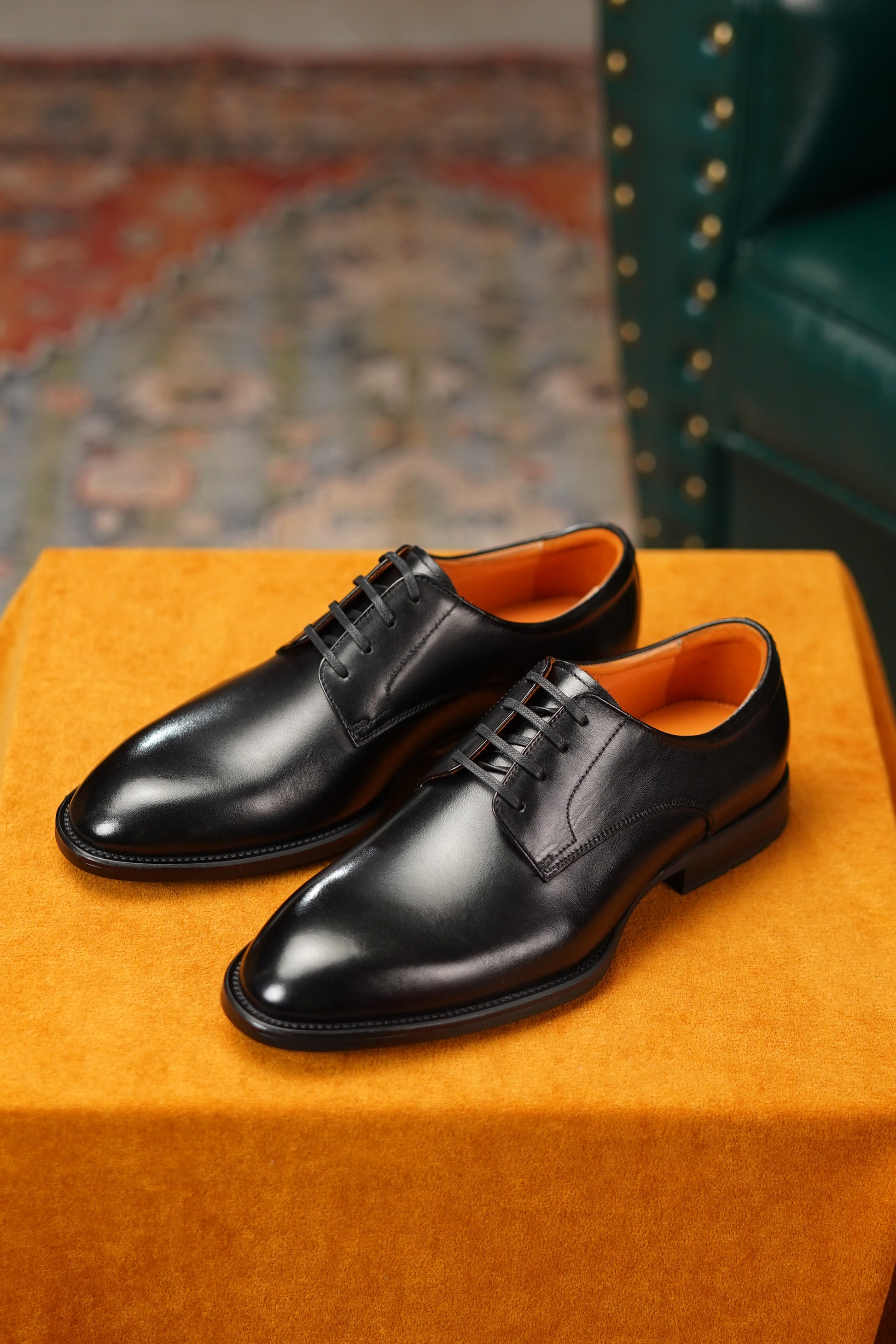 Pair of black dress shoes on an orange cushion with a patterned rug and green chair in the background.