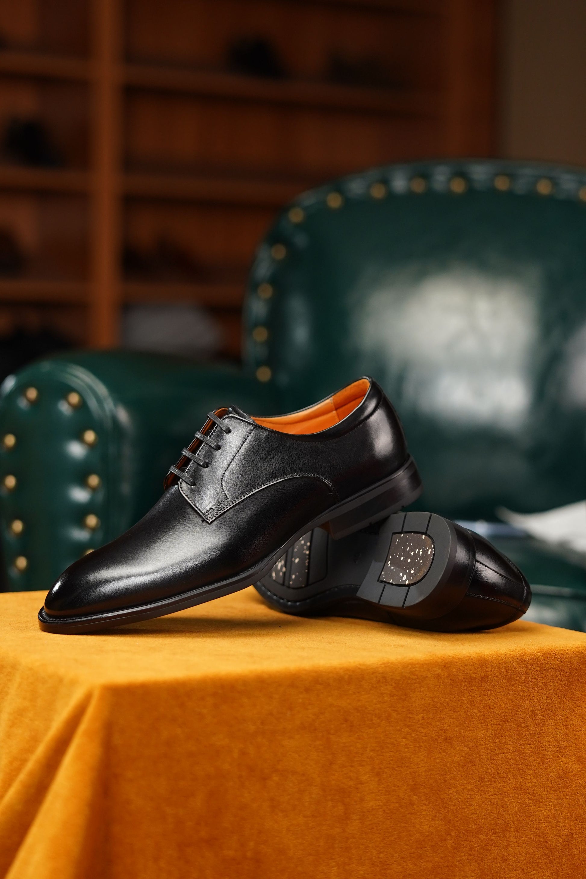 A pair of black leather oxford dress shoes with a contrasting orange interior lining, displayed on a draped mustard yellow fabric surface with a dark green leather armchair in the background.
