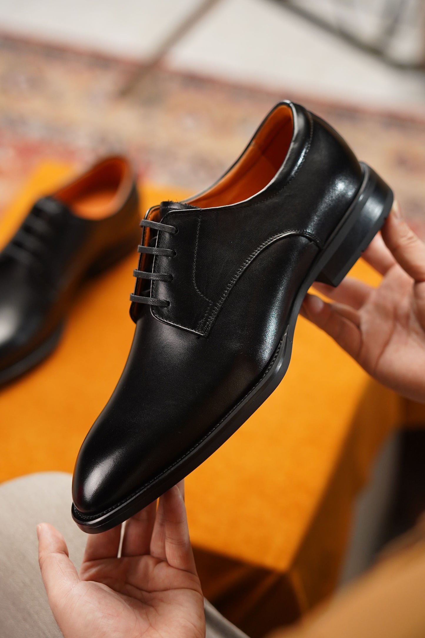 A man holds up a black leather oxford dress shoe with laces, showing the side profile and interior lining. Another matching black shoe is blurred in the background on an orange surface.