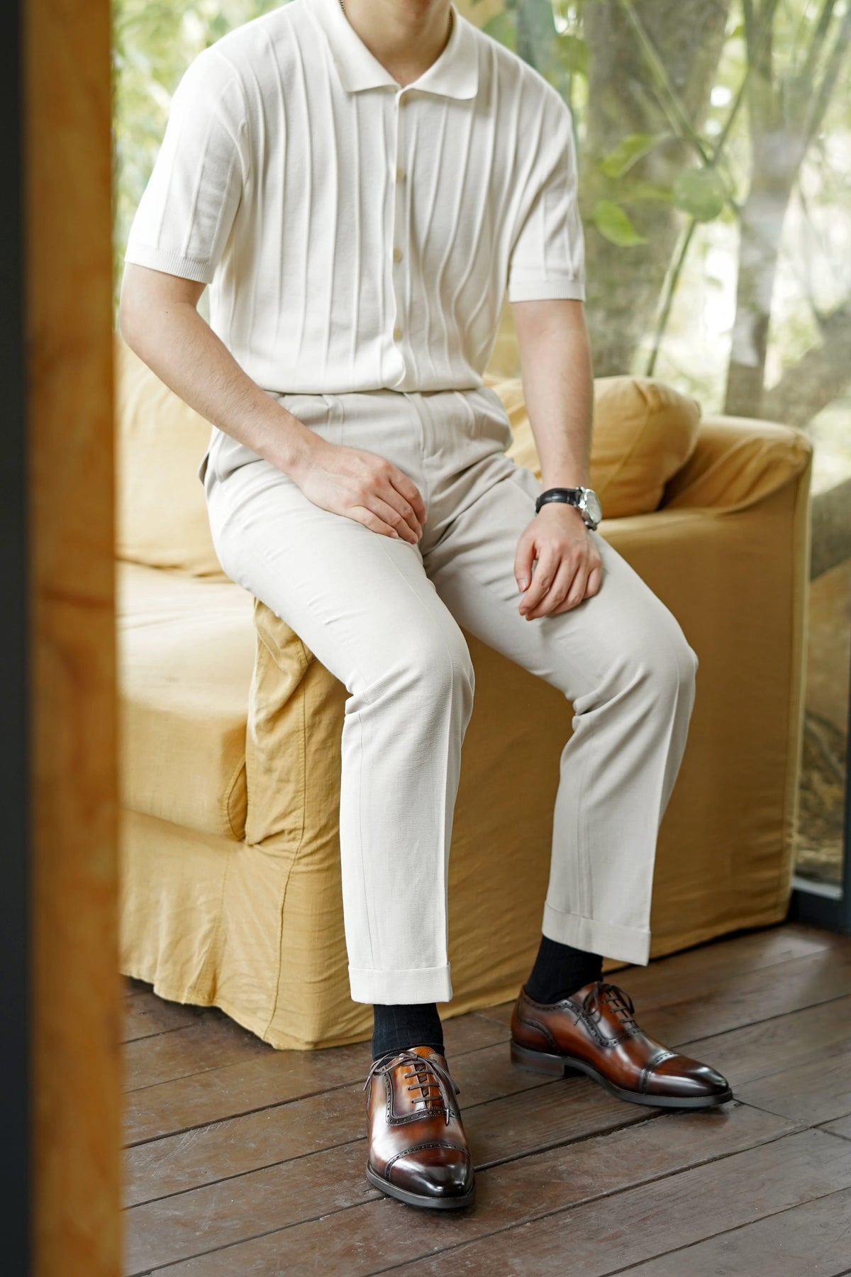 A man seated on a yellow sofa wearing a cream short-sleeve polo shirt, cream trousers, black socks, and brown oxford shoes in a warm indoor setting with a window and wooden floor.