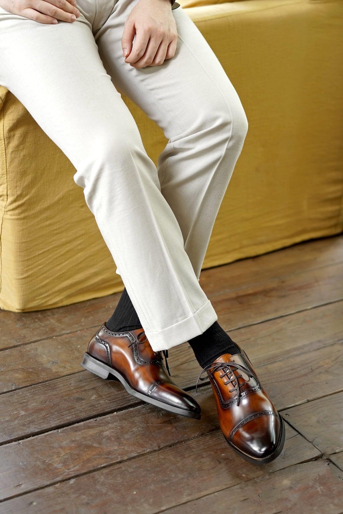 A close-up lifestyle shot of a person wearing brown leather brogue oxford shoes with black socks and cream trousers, sitting on a mustard yellow sofa over a wooden floor.