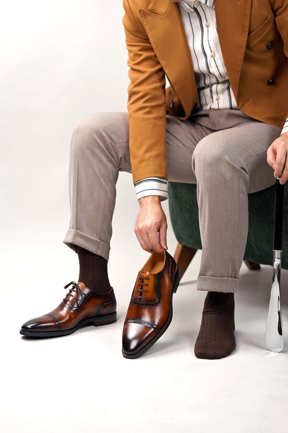 A man seated on a green stool wearing a tan blazer, striped shirt, beige trousers, dark socks, and brown leather oxford shoes, while holding up the third shoe to show the front. A golf club is visible on the right against a clean white studio background.