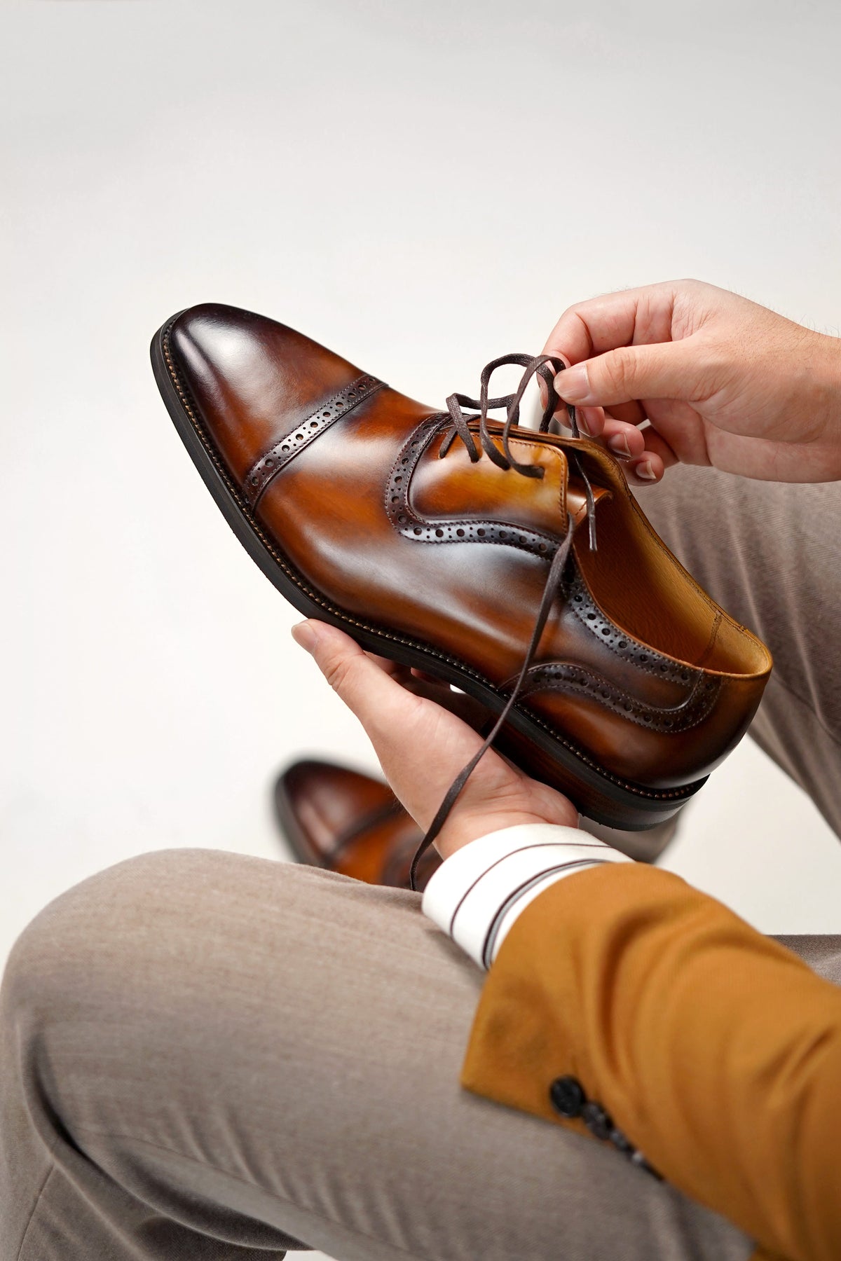 A person holds a polished brown leather wingtip shoe with dark laces, showing the toe and side profile. The person is wearing beige trousers and a mustard yellow jacket, with a second shoe blurred in the background against a light gray studio backdrop.
