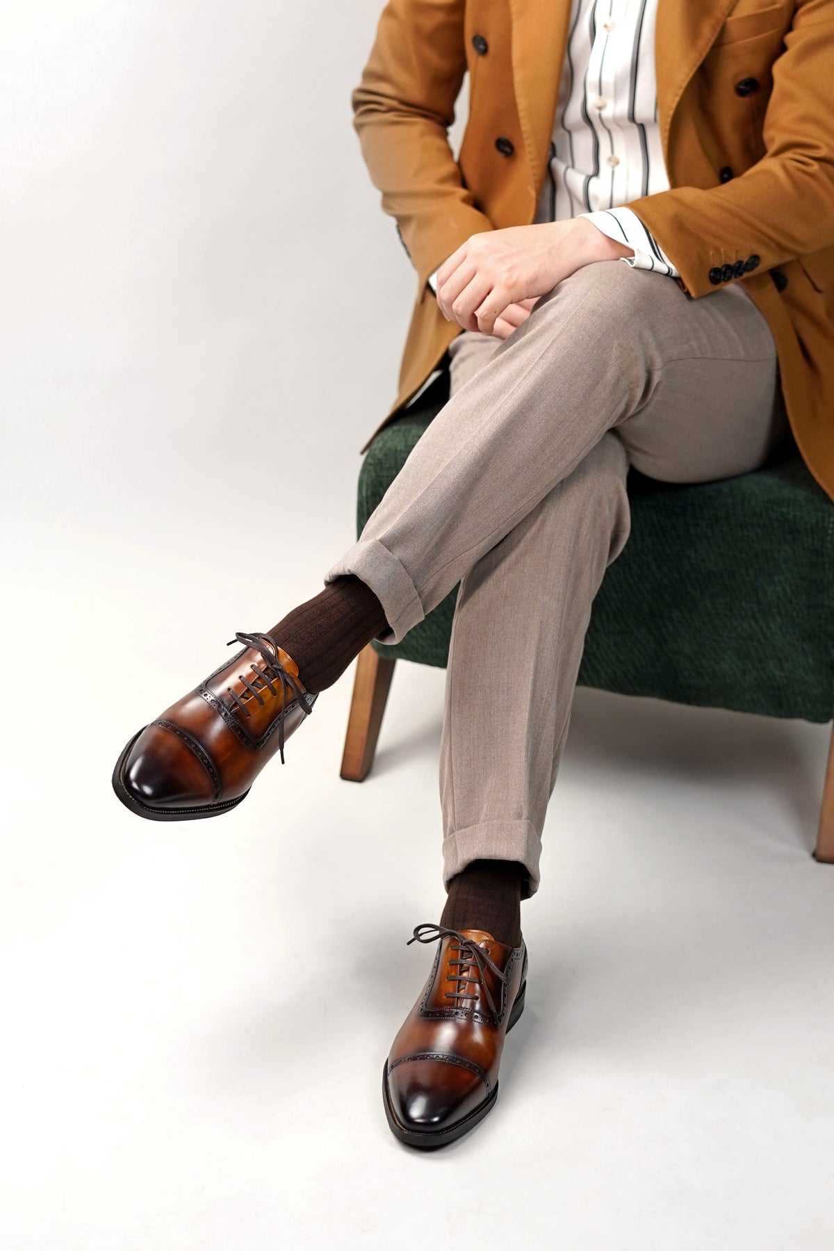 A person seated on a green upholstered chair wearing brown leather oxford shoes with a glossy toe cap, dark brown socks, light beige trousers, a white striped shirt, and a tan blazer against a light gray studio background.
