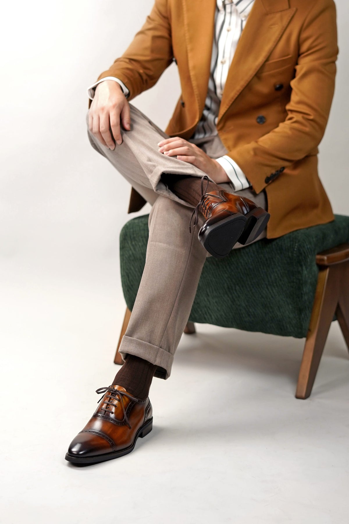 A man seated on a green upholstered chair wearing a camel brown double-breasted blazer, light beige trousers, dark brown dress shoes, and dark socks against a light gray studio background.