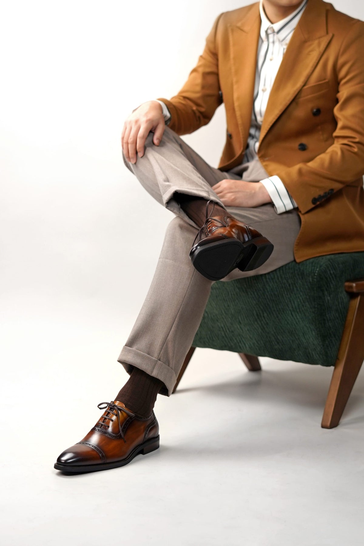 A man seated on a green upholstered chair wearing a camel brown double-breasted blazer, light gray trousers, dark brown dress socks, and brown oxford shoes with a black cap toe.