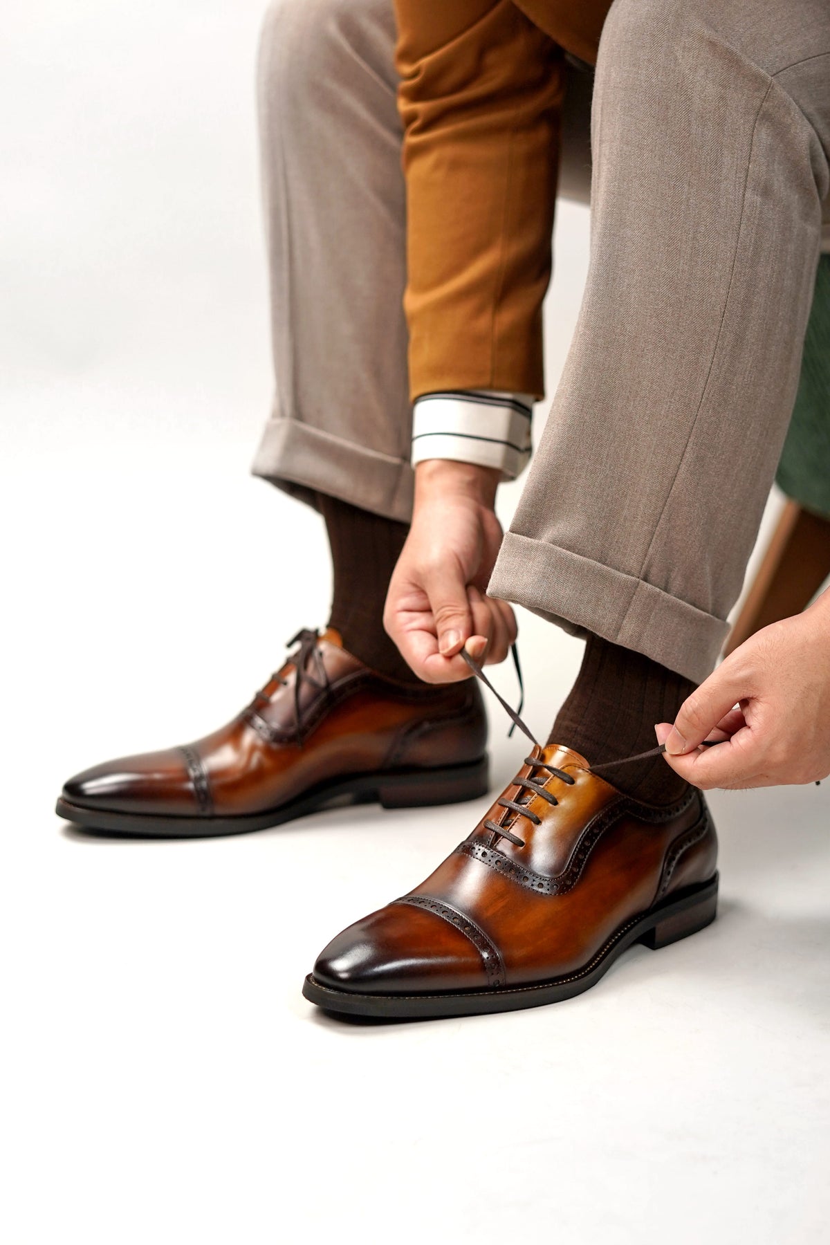 A close-up of a person wearing brown leather cap-toe oxford shoes with dark socks and light brown trousers, tying the laces of the front shoe against a clean white studio background.