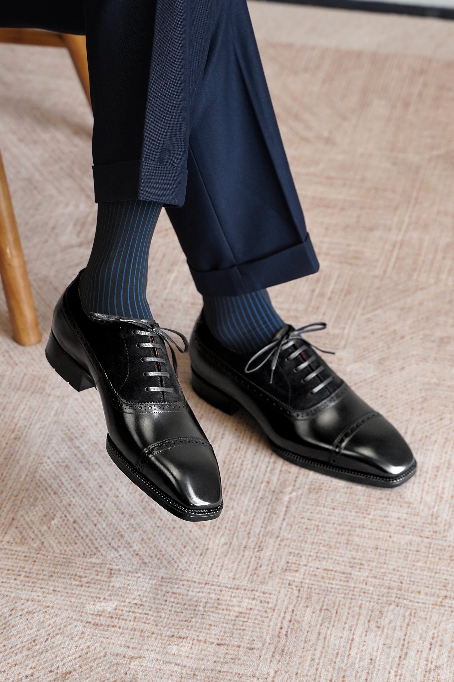 A person wearing black leather oxford dress shoes with dark blue and navy striped socks and navy trousers, standing on a textured beige carpet.