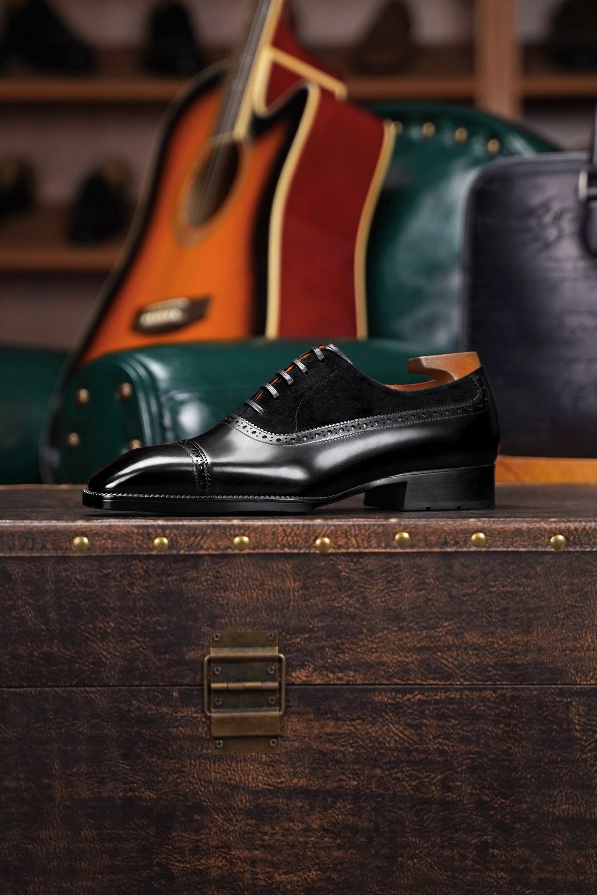 A black leather cap-toe oxford shoe with brogue detailing, displayed on a dark wooden trunk with an acoustic guitar and leather goods in the background.