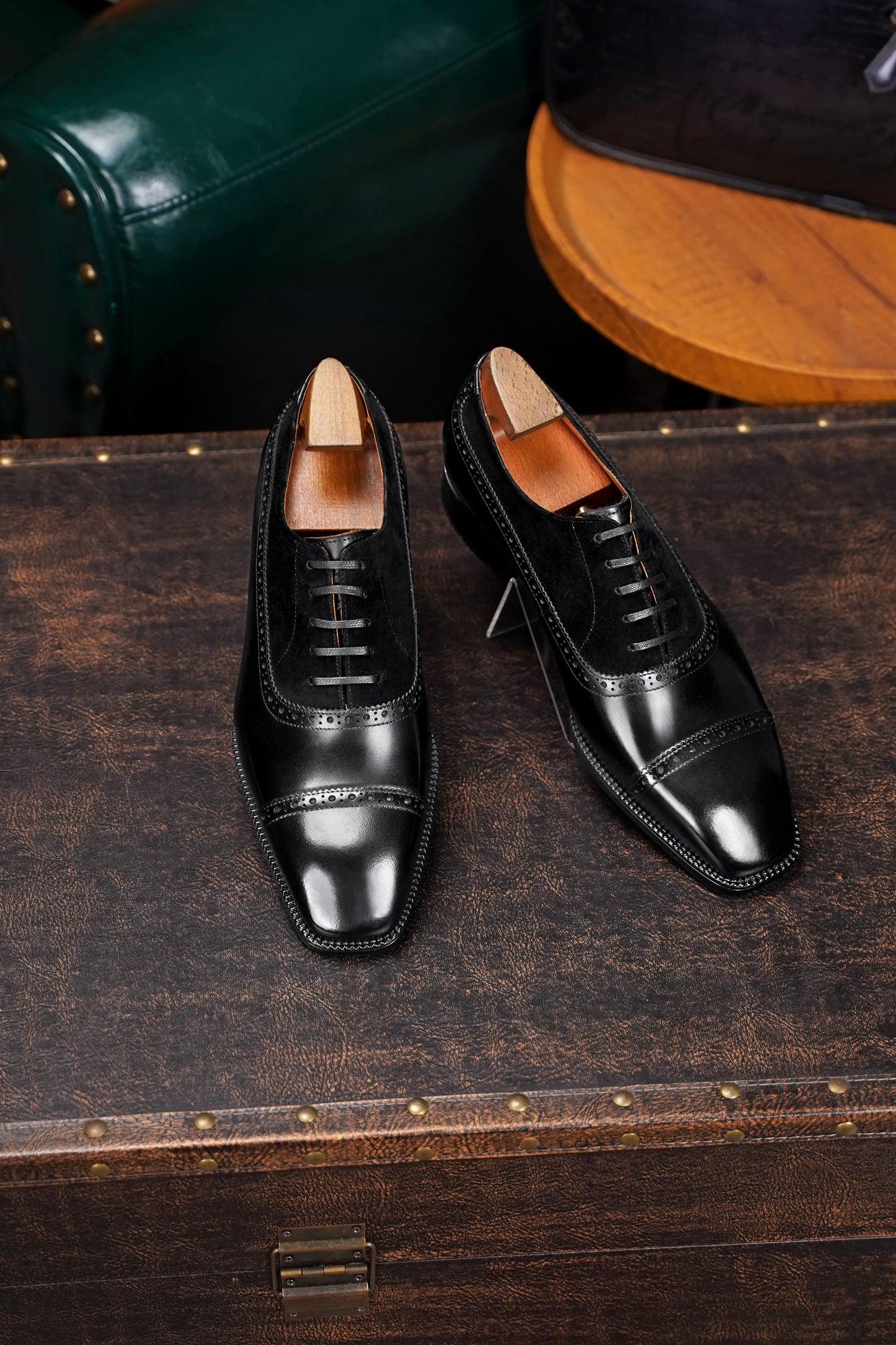 Pair of black dress shoes on a wooden surface with a dark green leather chair in the background.