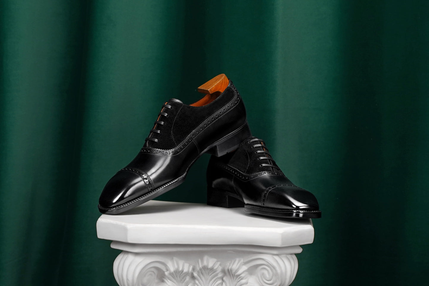 Pair of black dress shoes on a white pedestal against a green curtain background