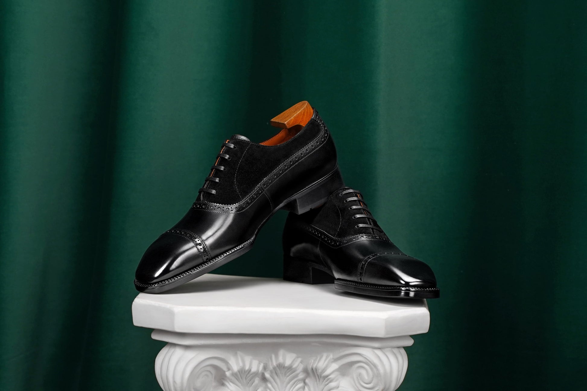 Pair of black dress shoes on a white pedestal against a green curtain background