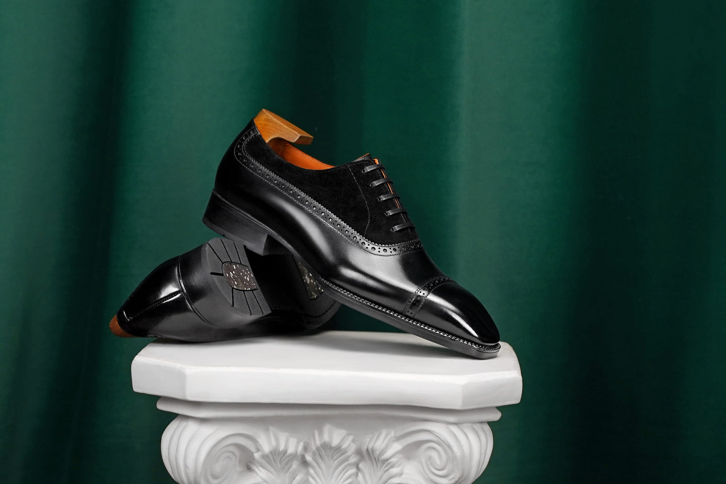 A pair of black leather cap-toe oxford shoes with brogue detailing and orange heel inserts, displayed on a white decorative pedestal against a dark green curtain background.
