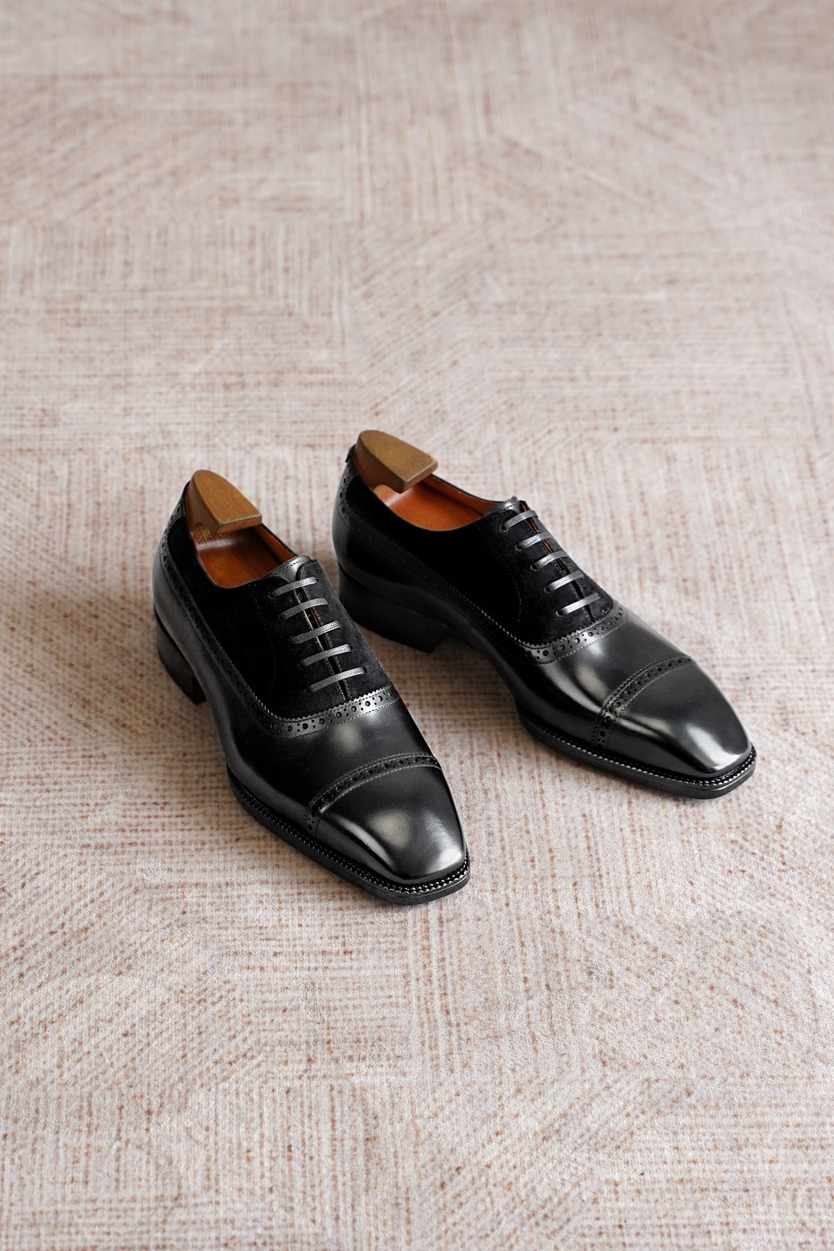 A pair of black leather cap-toe oxford shoes with brogue detailing, displayed on a textured beige woven rug.