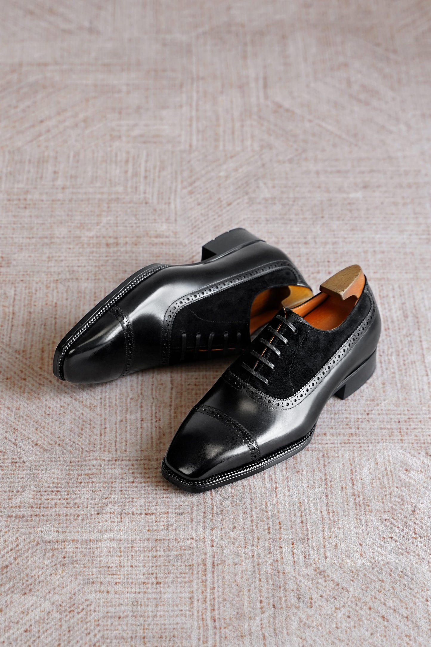 A pair of men's black leather wingtip oxford shoes with suede detailing, displayed on a textured beige rug.