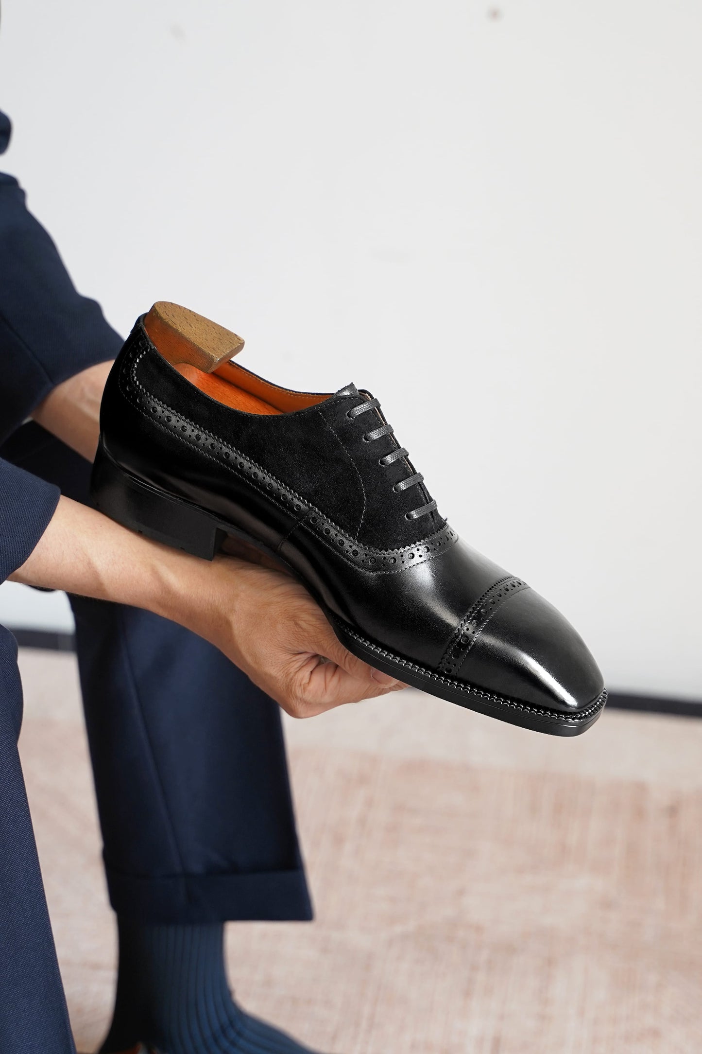 A person holds up a black leather cap-toe oxford shoe with brogue detailing, showing the orange lining and wooden shoe tree insert, against a neutral indoor background.