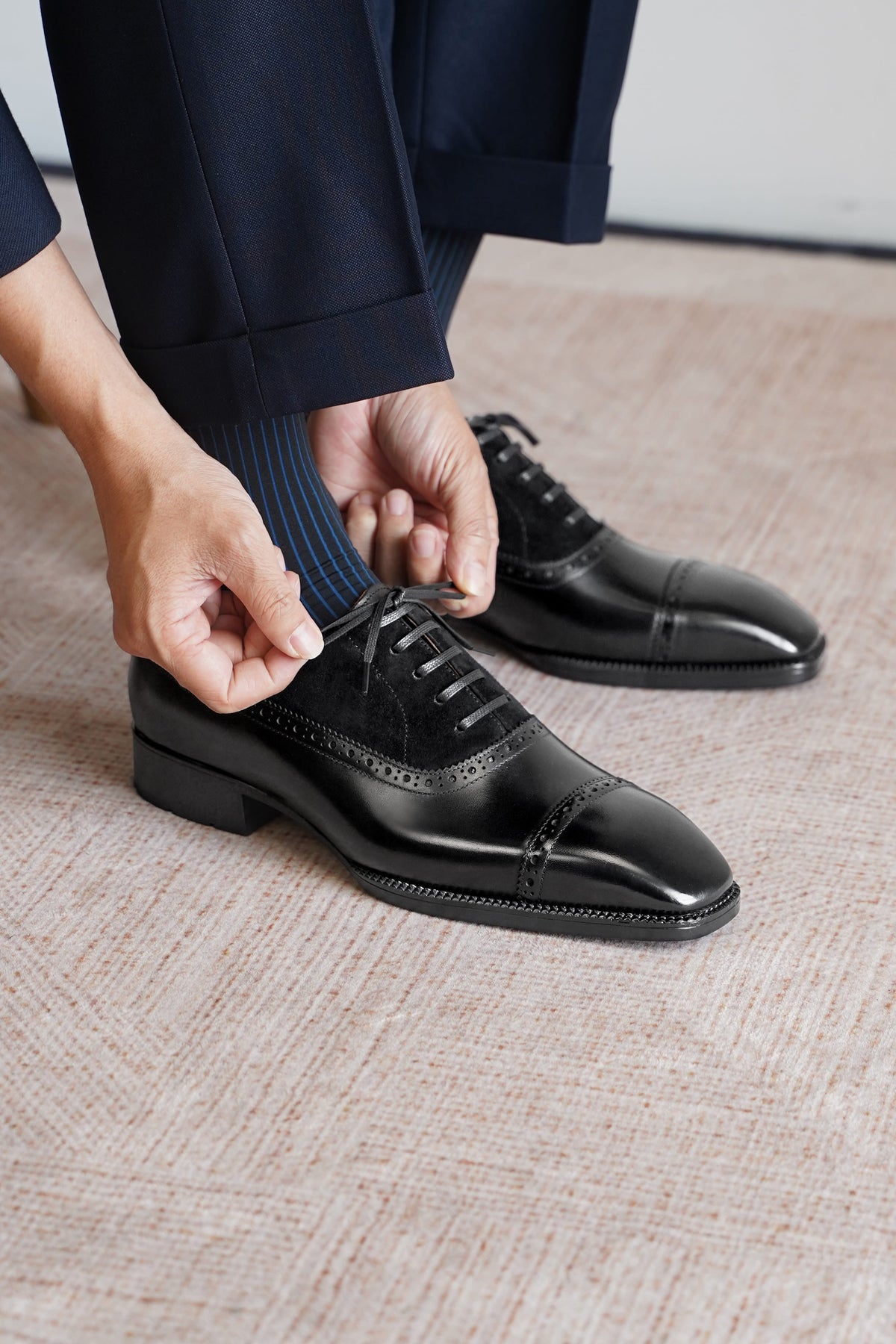 A close-up of a person's hands tying the laces of a black leather cap-toe oxford shoe, with navy trousers and striped socks visible, set on a textured beige carpet.