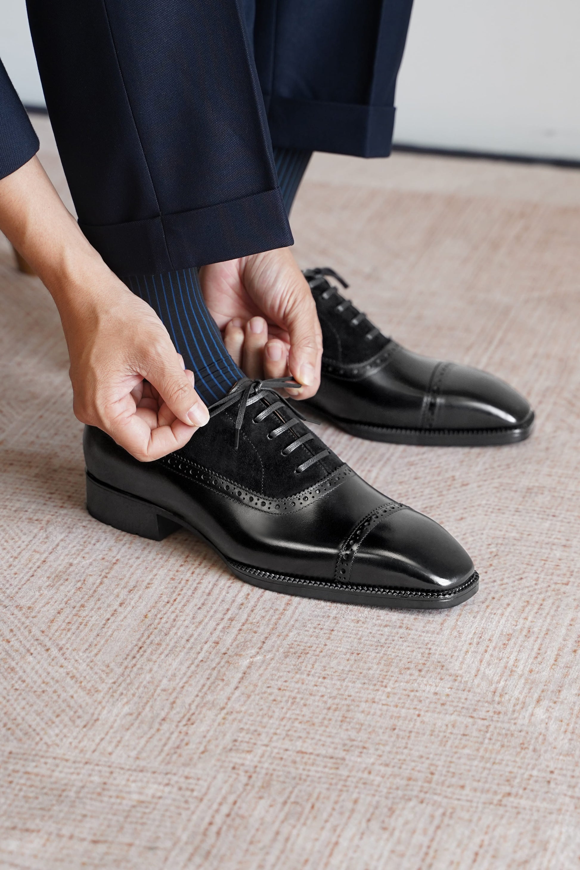 A close-up of a person's hands tying the laces of a black leather cap-toe oxford shoe, with navy trousers and striped socks visible, set on a textured beige carpet.