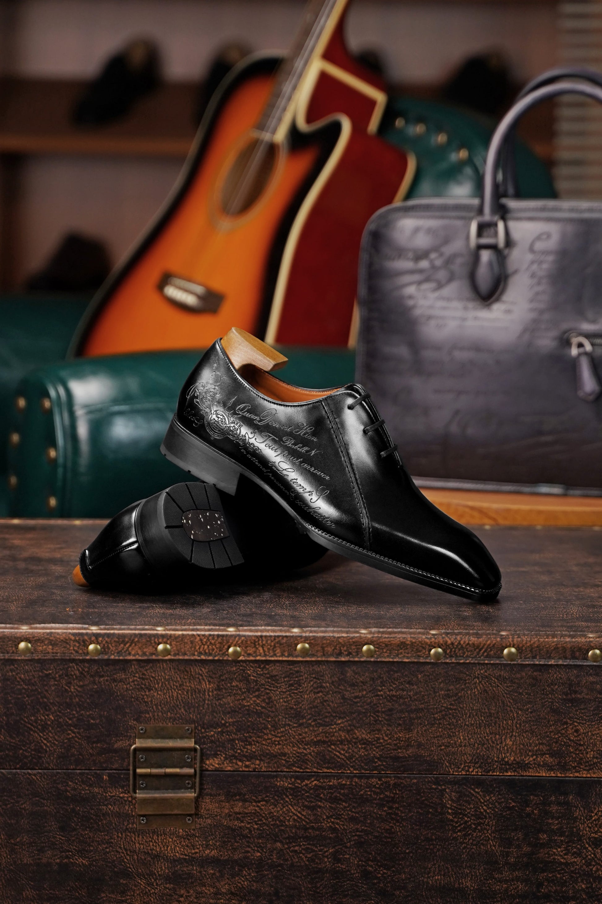 A pair of black leather dress shoes with a glossy finish and subtle grain texture, displayed on a vintage wooden trunk. The background features a dark green leather armchair, an acoustic guitar, and a textured gray handbag.