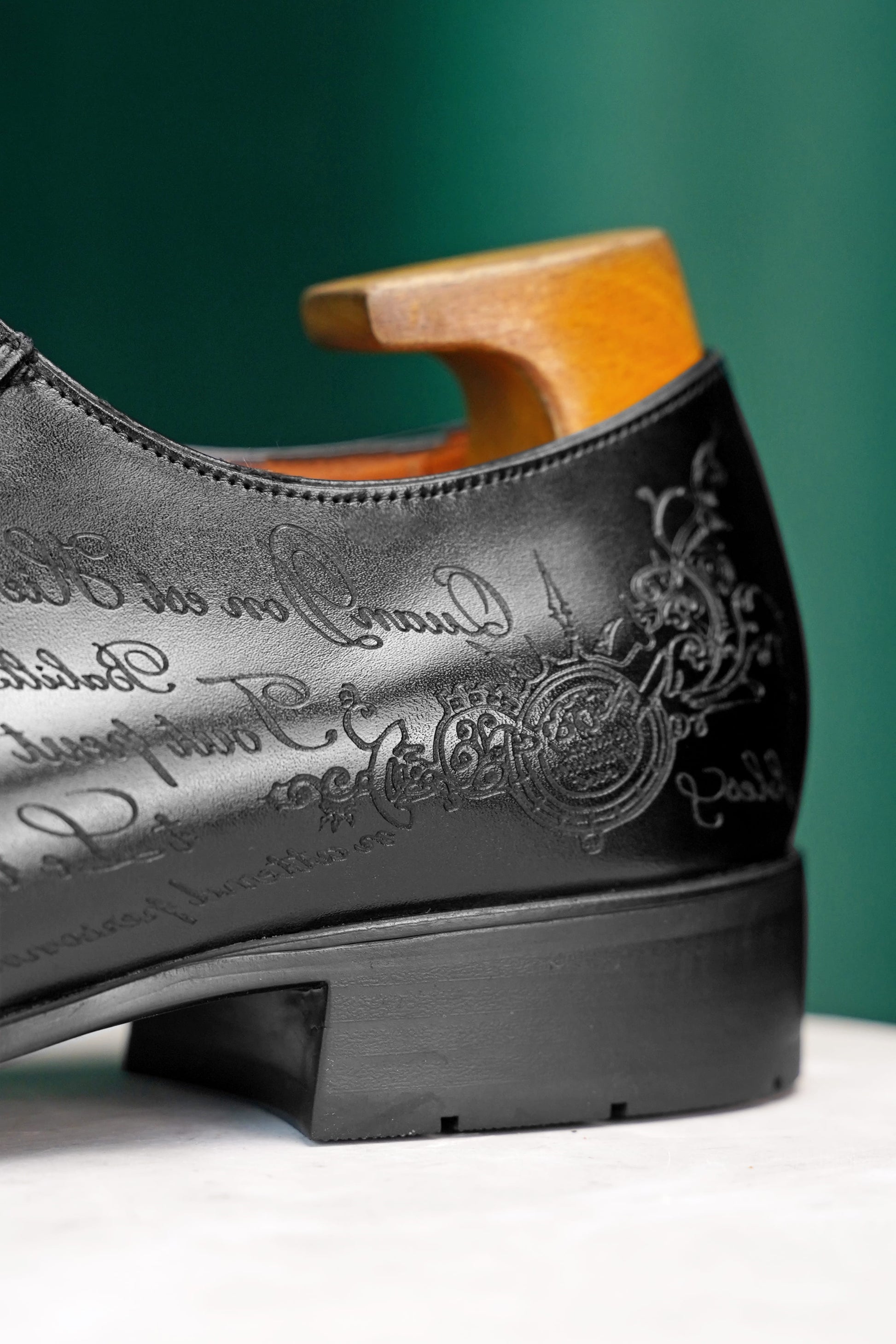 A close-up of a black leather dress shoe with embossed script and crest detailing, shown with a wooden shoe tree insert against a dark green background.