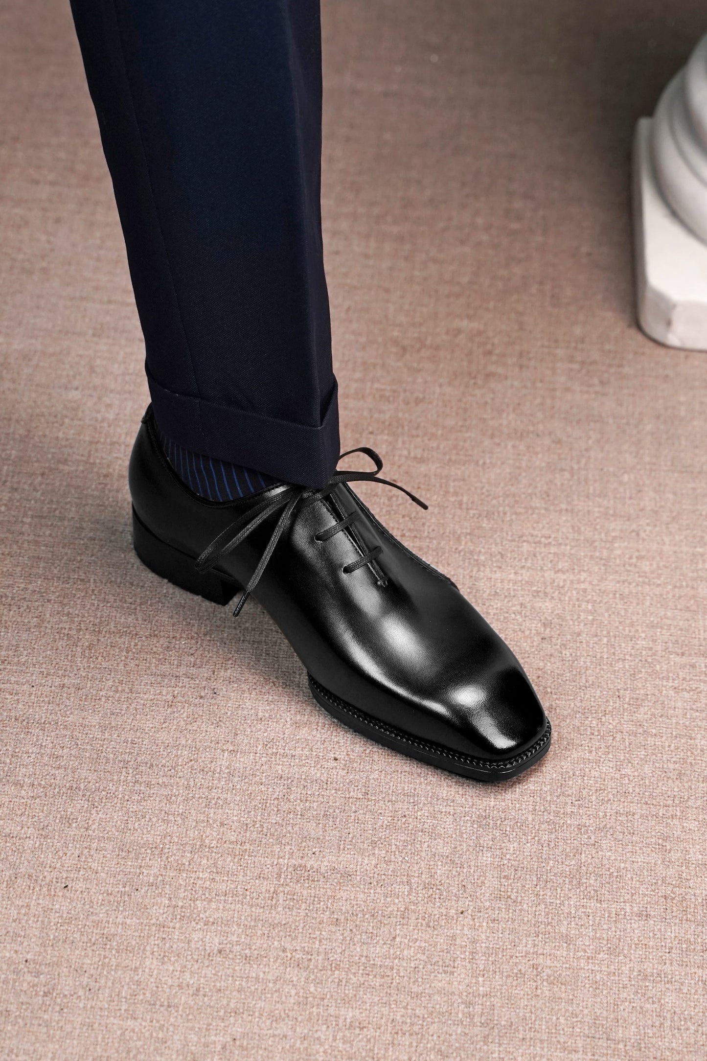 A close-up of a man's foot wearing a polished black leather oxford dress shoe with dark navy trousers and a striped sock, standing on a textured beige carpet.