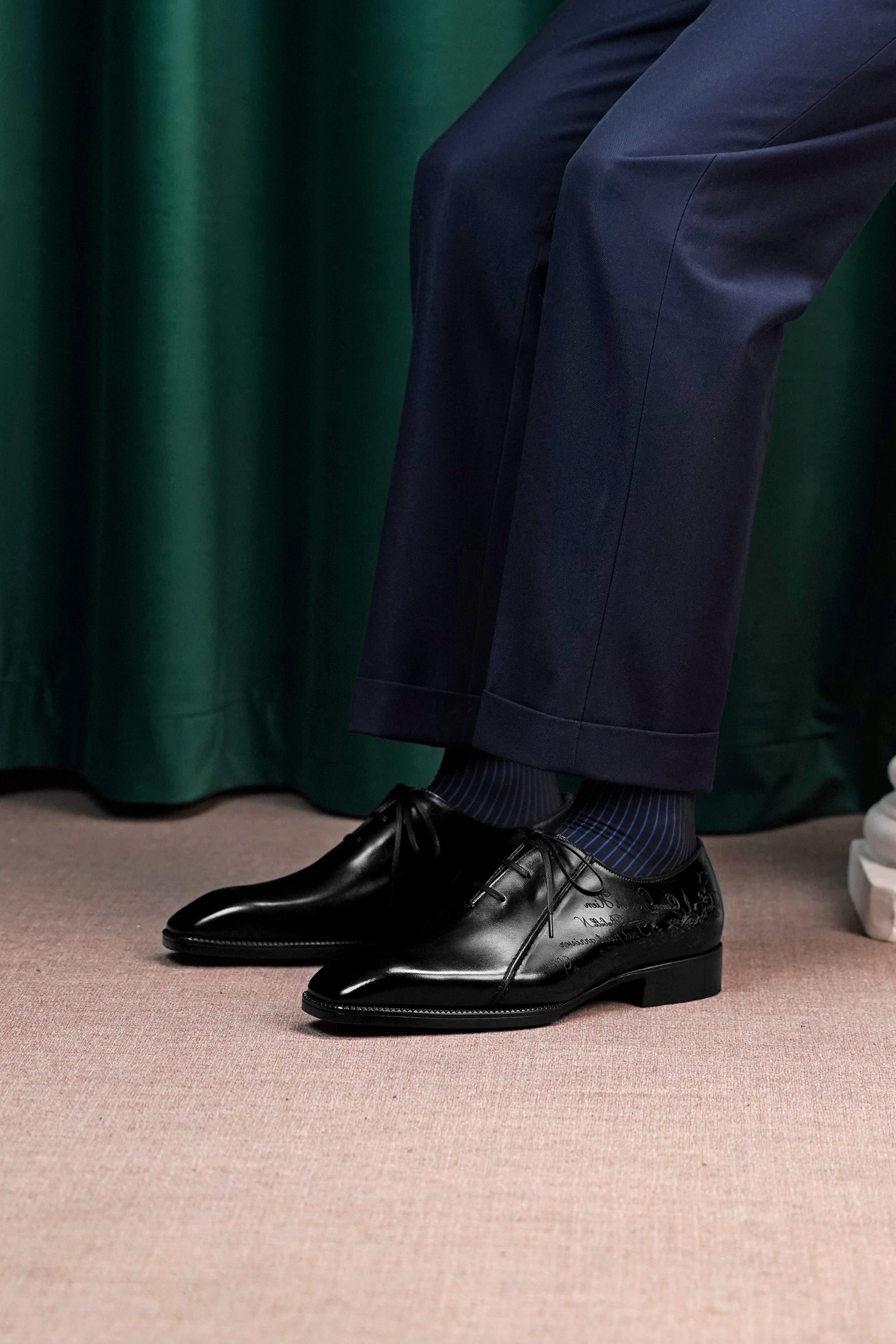 A close-up of a person's legs wearing navy blue trousers, dark blue socks, and shiny black leather oxford dress shoes, set against a dark green curtain and beige carpet.