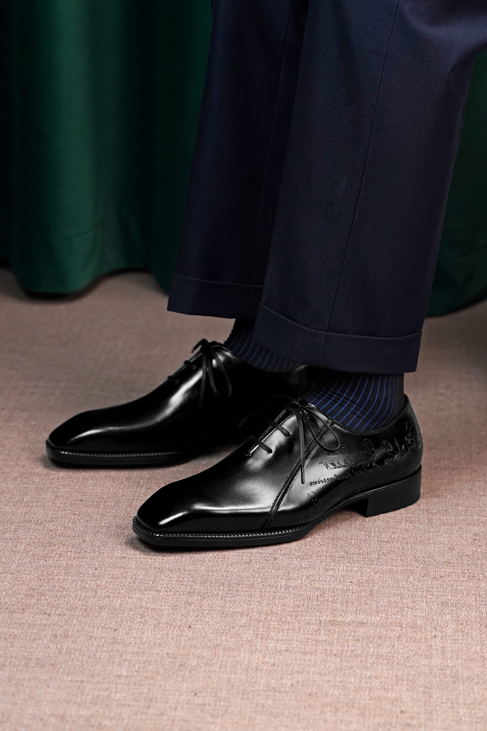 A close-up of a person wearing polished black lace-up dress shoes with navy blue socks and navy trousers, standing on a textured beige floor with a dark green curtain in the background.