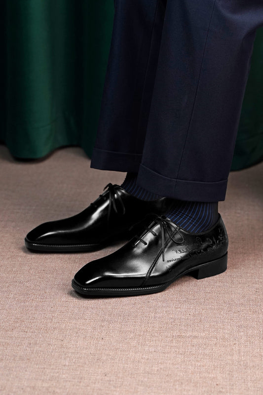 A close-up of a person wearing polished black lace-up dress shoes with navy blue socks and navy trousers, standing on a textured beige floor with a dark green curtain in the background.