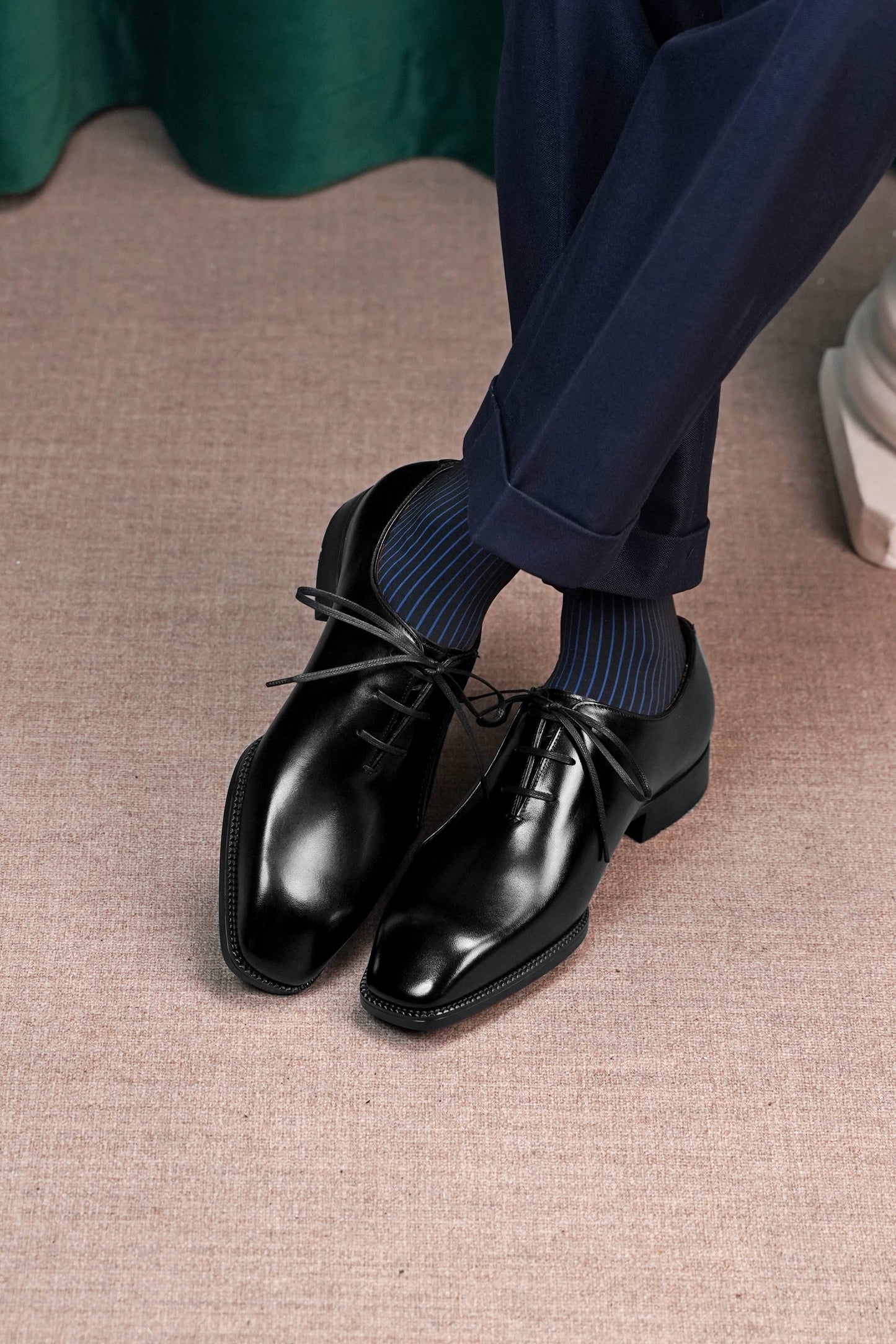 A pair of men's black leather oxford dress shoes with a pointed toe, worn with navy blue trousers and dark blue striped socks on a textured beige floor.