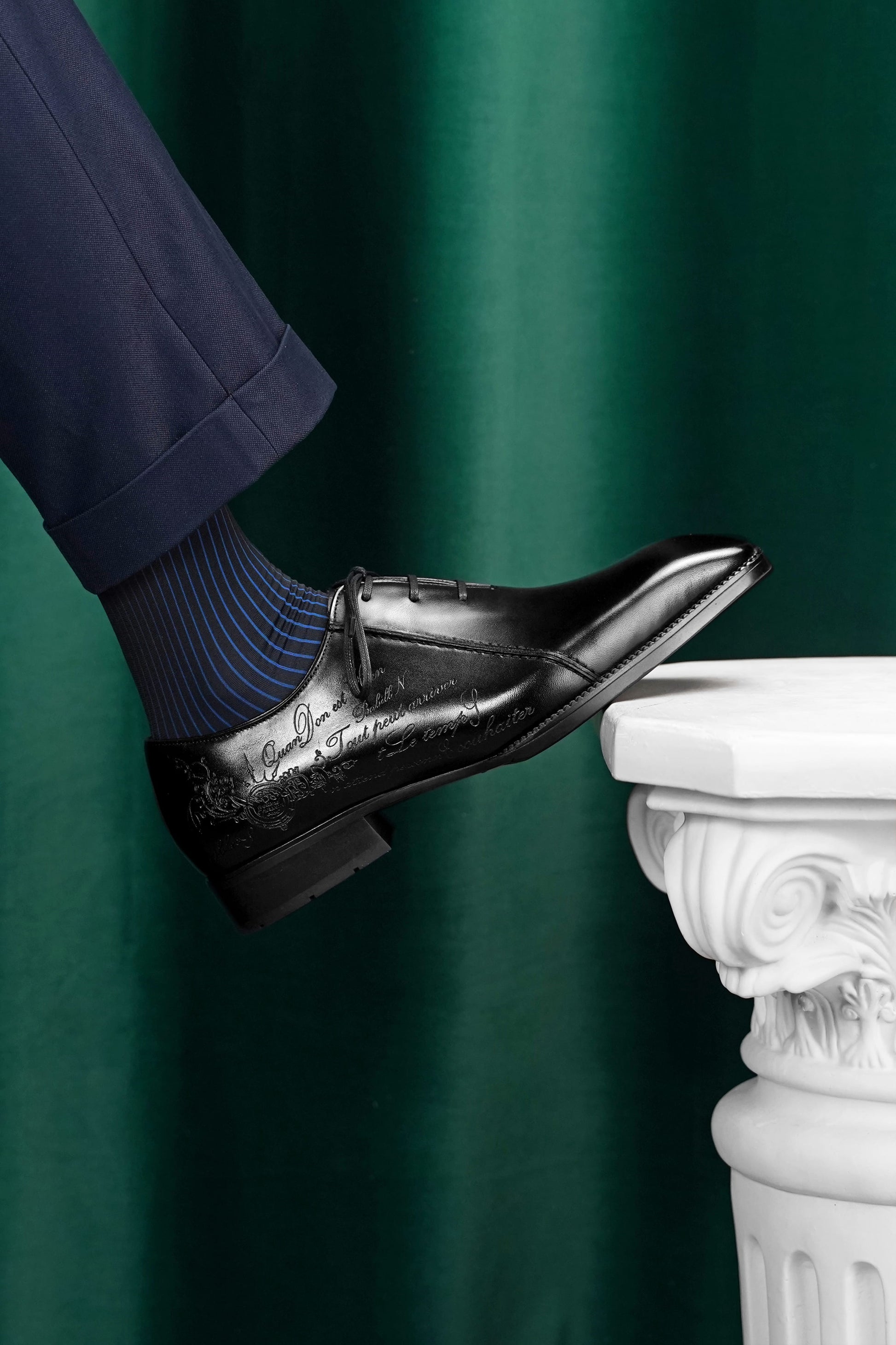 A man's foot wearing a black embossed oxford dress shoe with a blue and navy striped sock, resting on a white classical column against a dark green curtain backdrop.