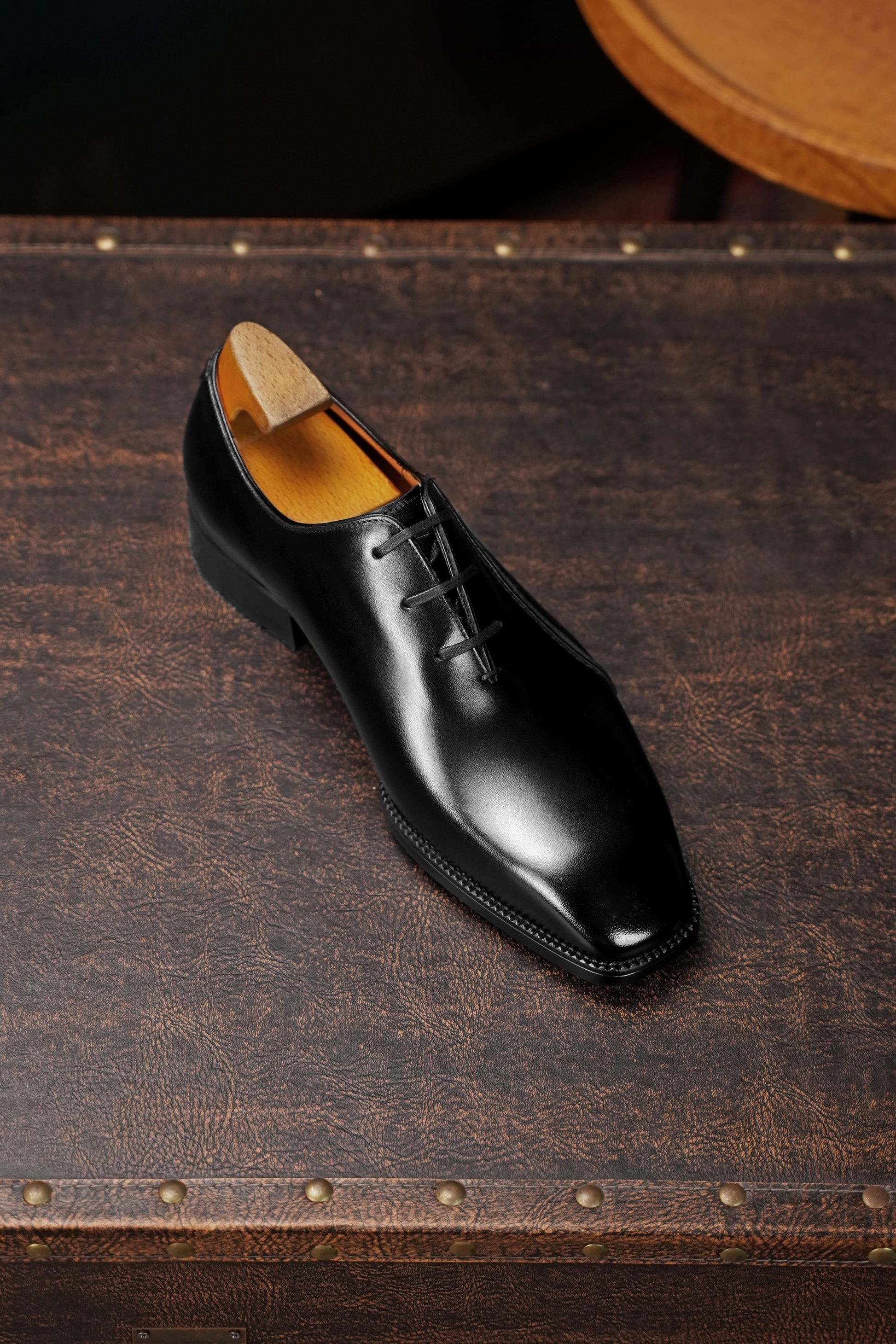 A single black leather dress shoe with a pointed toe and lace-up front, displayed on a dark brown textured leather surface.