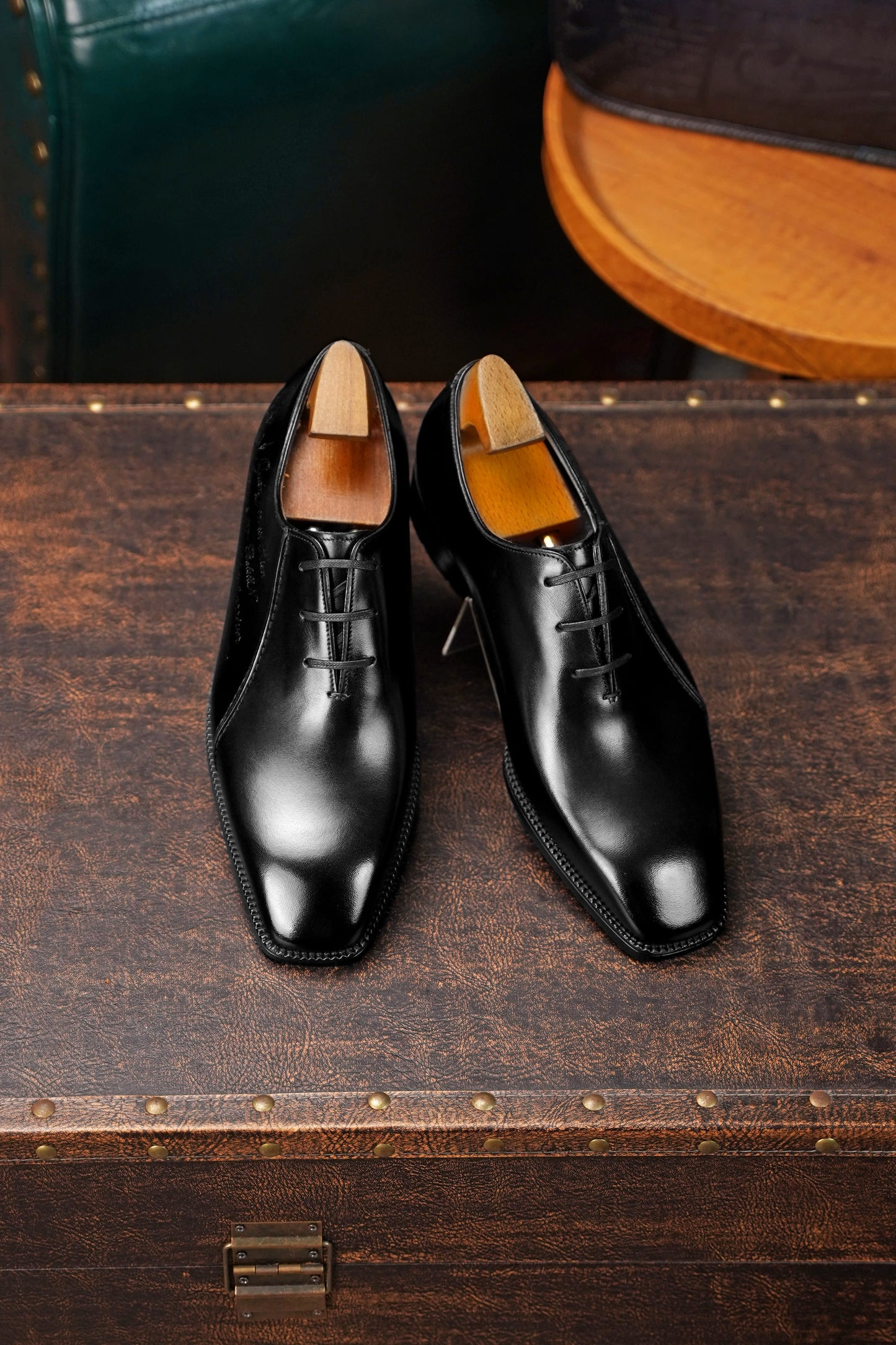 A pair of polished black leather oxford dress shoes with black laces and light tan insoles, displayed on a rustic dark brown wooden trunk.