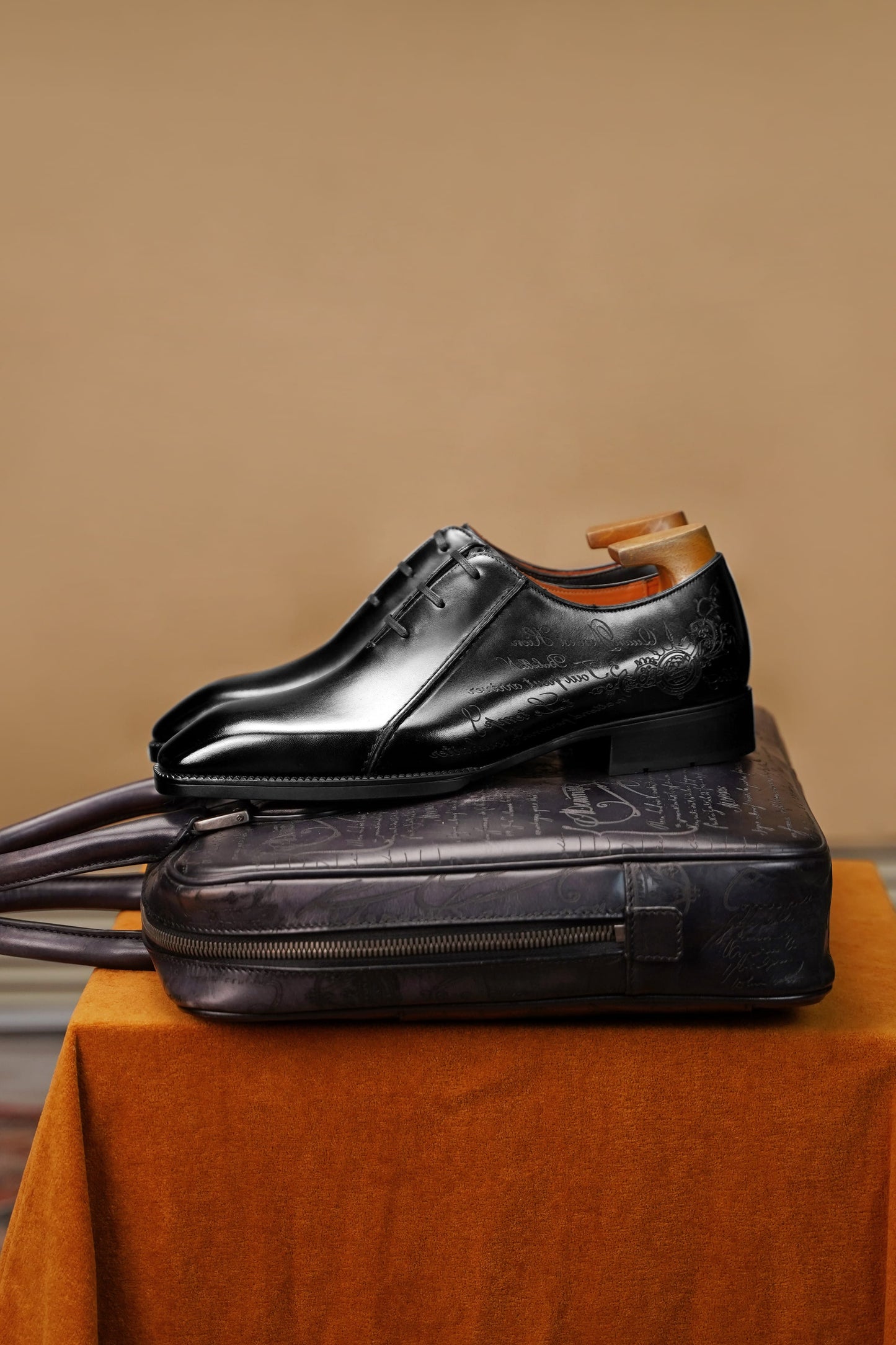A pair of black leather dress shoes with wooden shoe trees, displayed on top of a dark leather case resting on an orange fabric-covered pedestal against a warm beige background.
