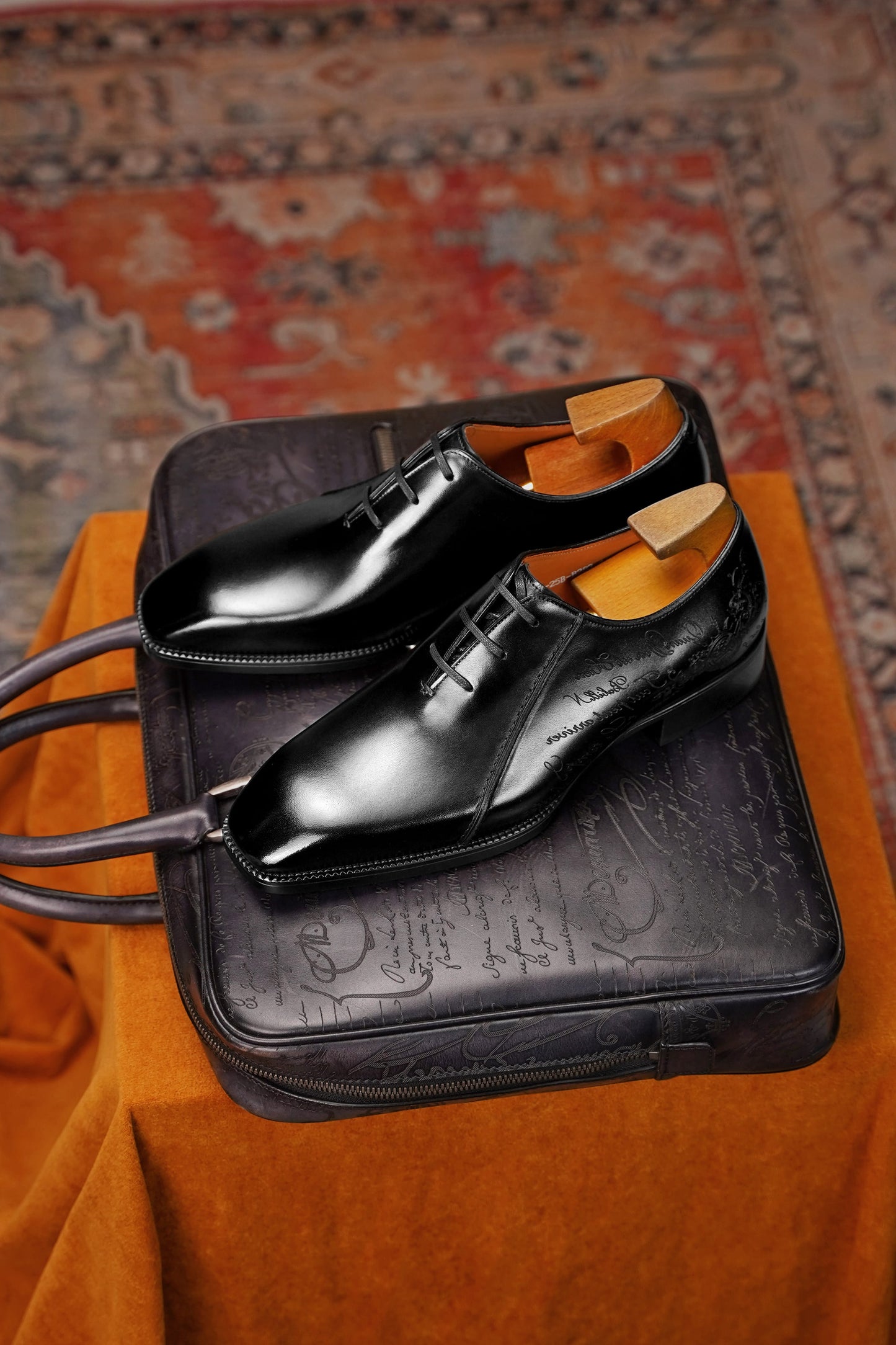 A pair of polished black leather oxford dress shoes with light wood shoe trees, displayed on top of a black embossed leather briefcase on an orange fabric surface with a patterned rug in the background.